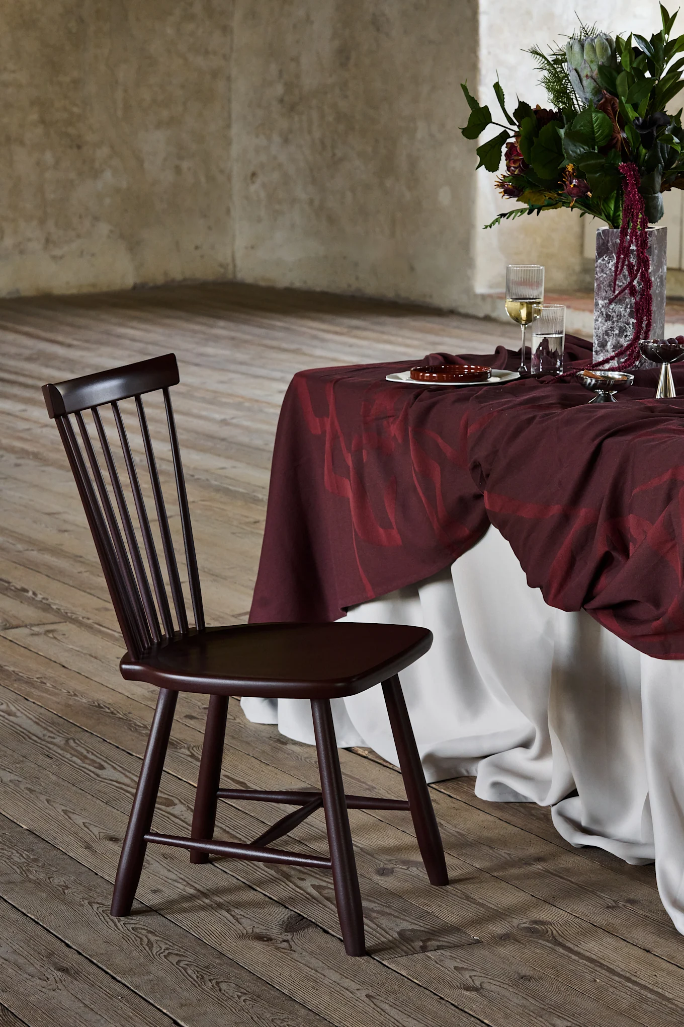 Dark brown wooden chair with slatted back, next to a table draped in burgundy with a floral arrangement, on a wooden floor.