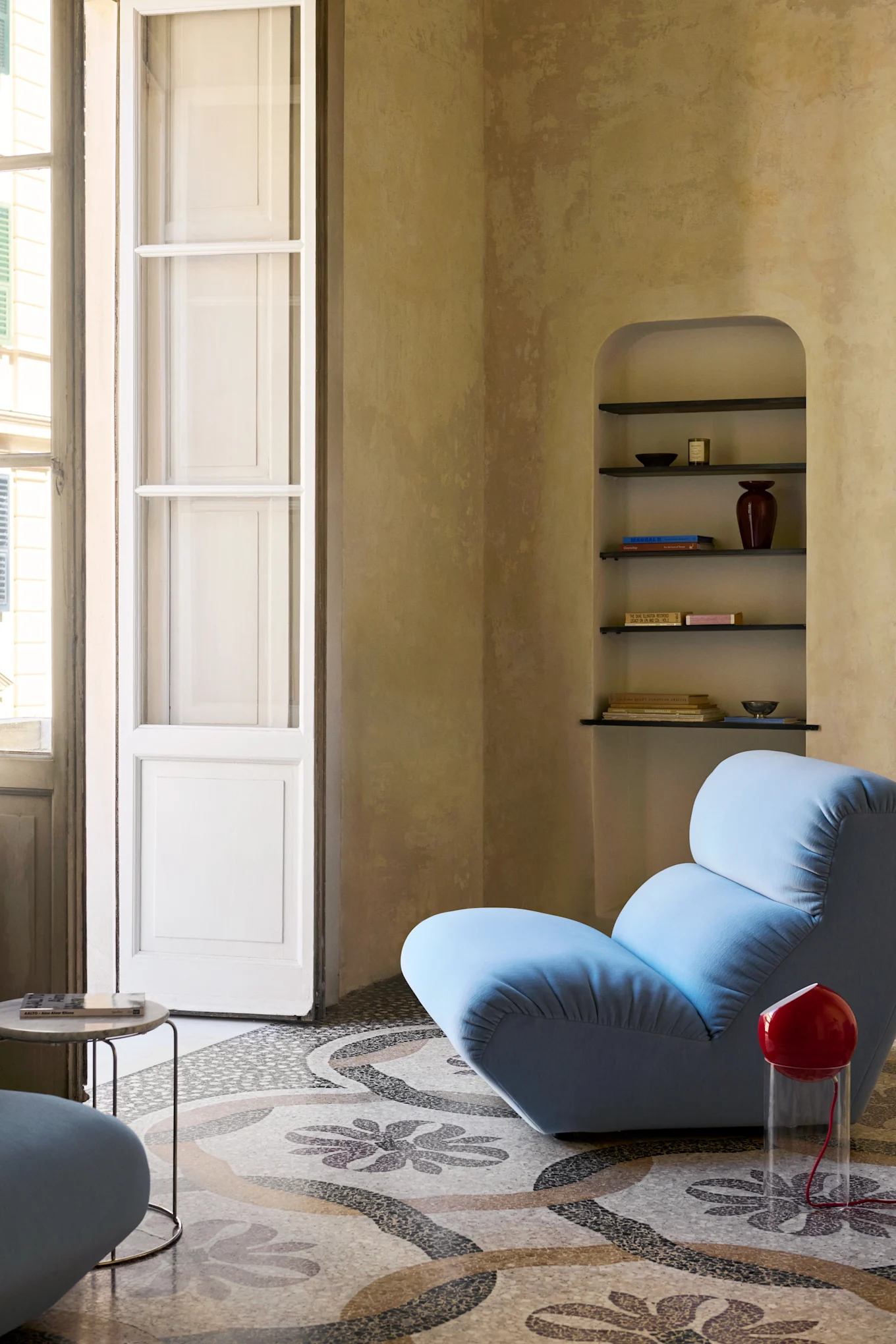 A light blue lounge chair on a patterned mosaic floor next to a red lamp, with an arched shelf and tall window.