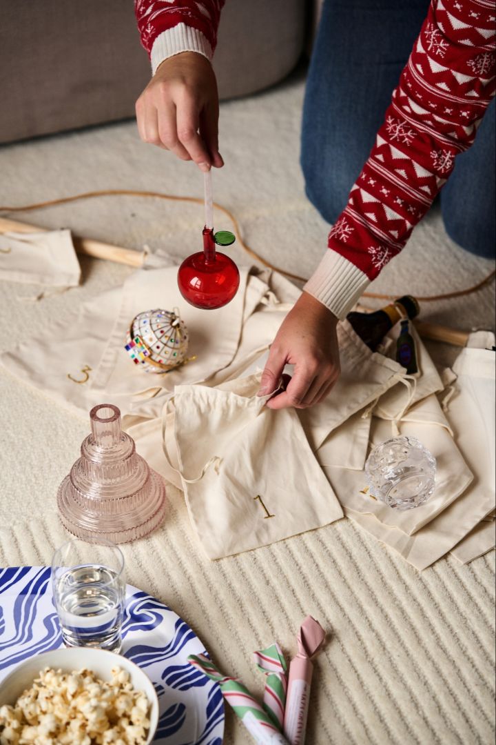 Hands place a glass red apple decoration from Iittala into the bag of an Advent calendar from ferm LIVING. 
