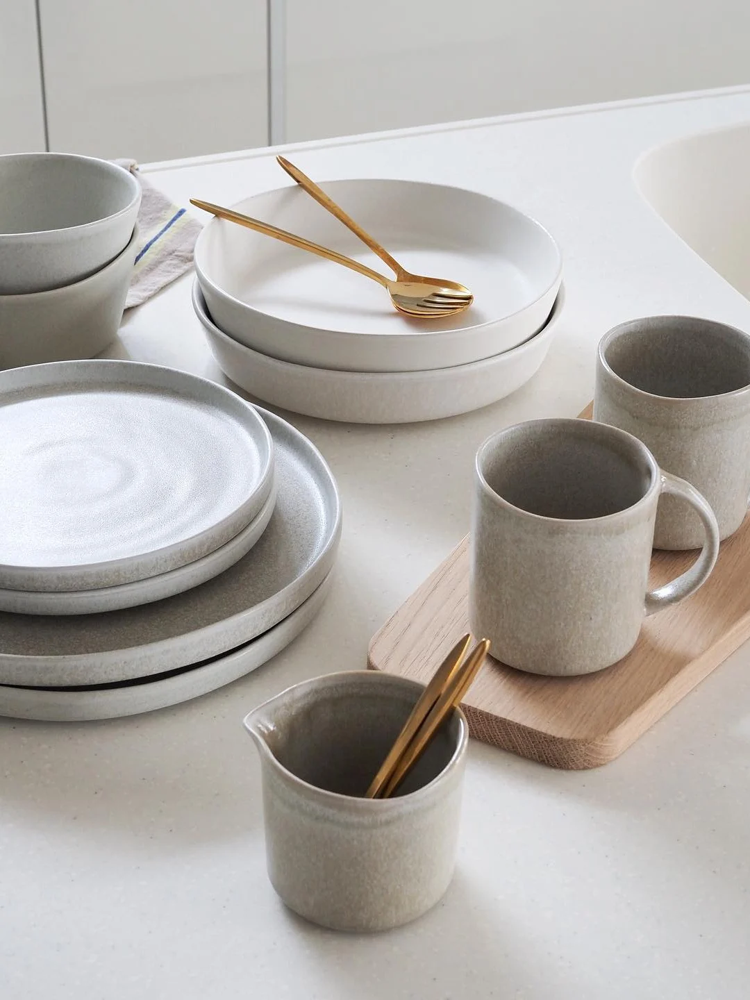 Modern gray and white ceramic dinnerware, including stacked plates, bowls, and mugs, with gold utensils on a kitchen counter.