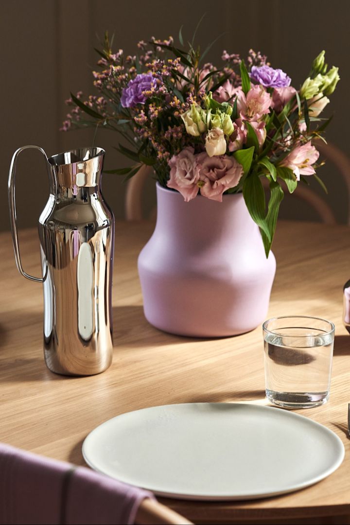 Here you can see the purple Dorotea vase with a bouquet of pink and purple flowers standing on a table next to the silver Dorotea carafe.