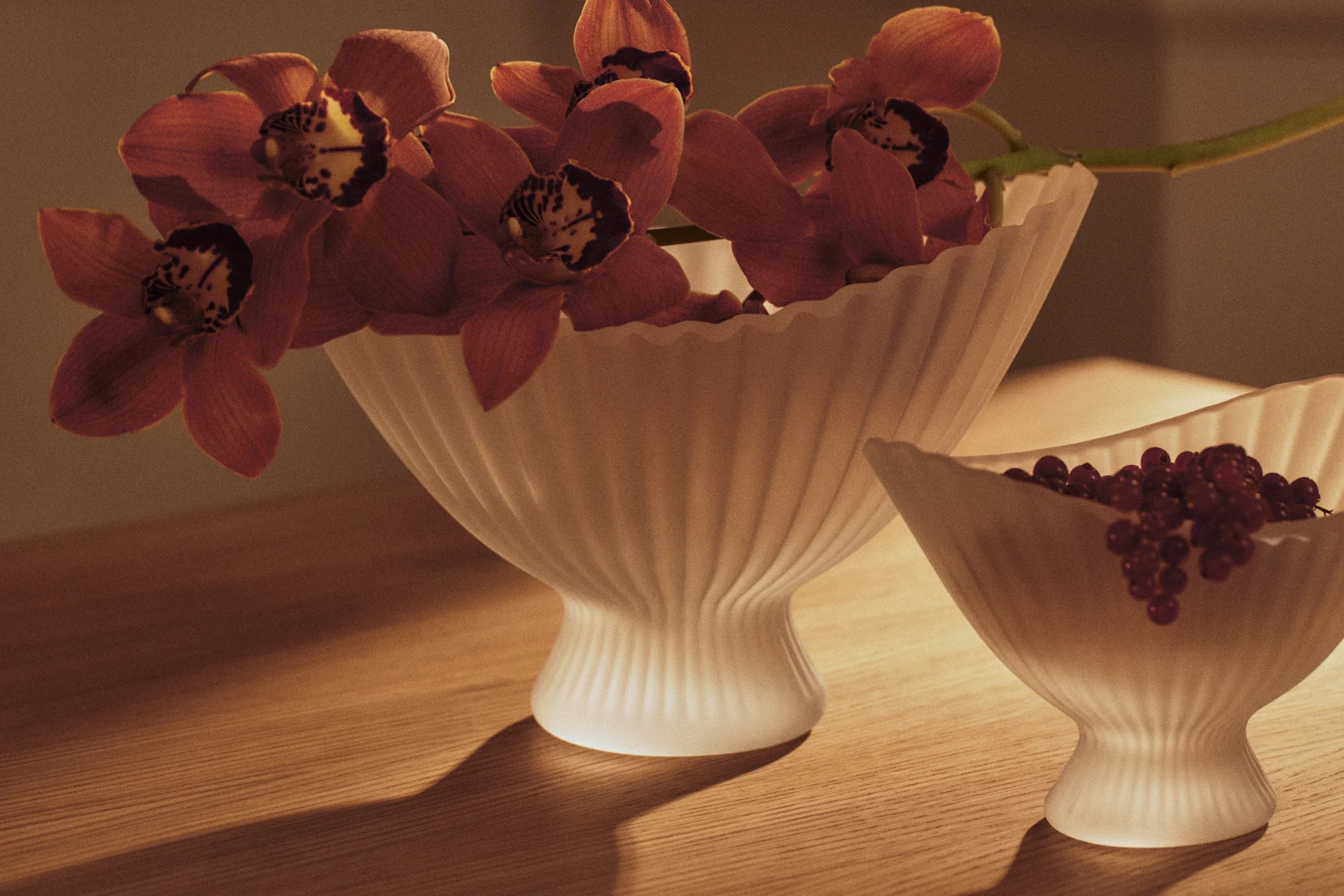 Red-orange orchids in a fluted glass vase and red currants in a smaller matching bowl on a wooden table.