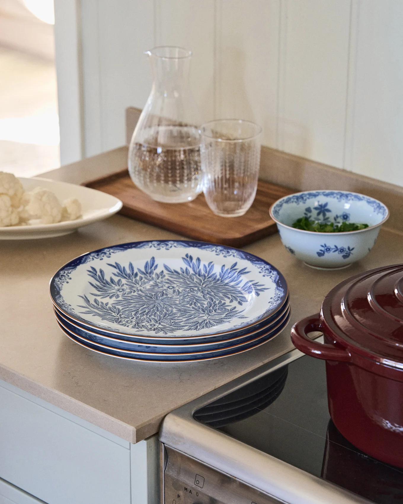 Stacked white plates with blue floral pattern, next to a water carafe, a bowl of herbs, and a red pot on a kitchen counter.