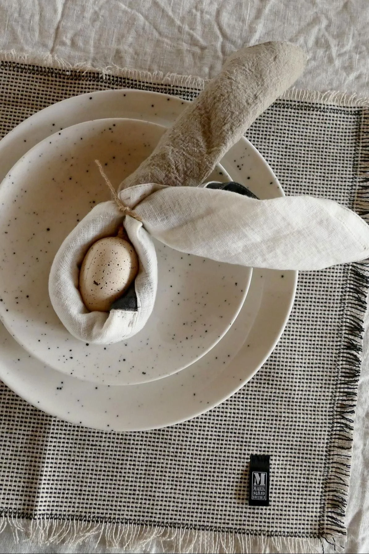 Easter table setting with speckled plates, linen napkins folded as bunny ears holding a speckled egg, and a checkered placemat.