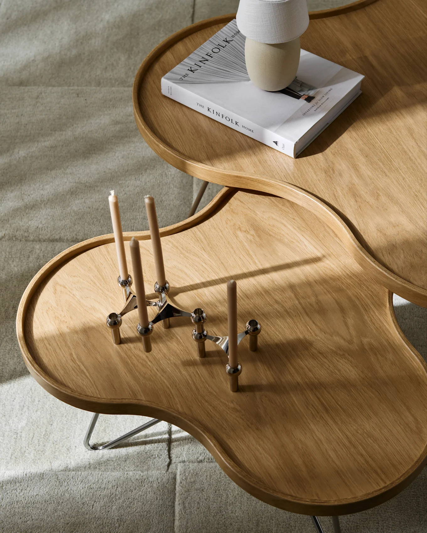 Two irregular wooden coffee tables on a light grey carpet. One table has a book and a lamp, the other has two candle holders with candles.