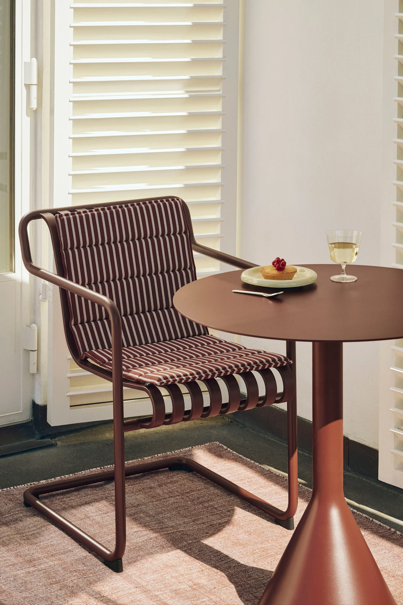 A reddish-brown chair with a striped cushion next to a matching round table holding a cake with berries and a glass of white wine.
