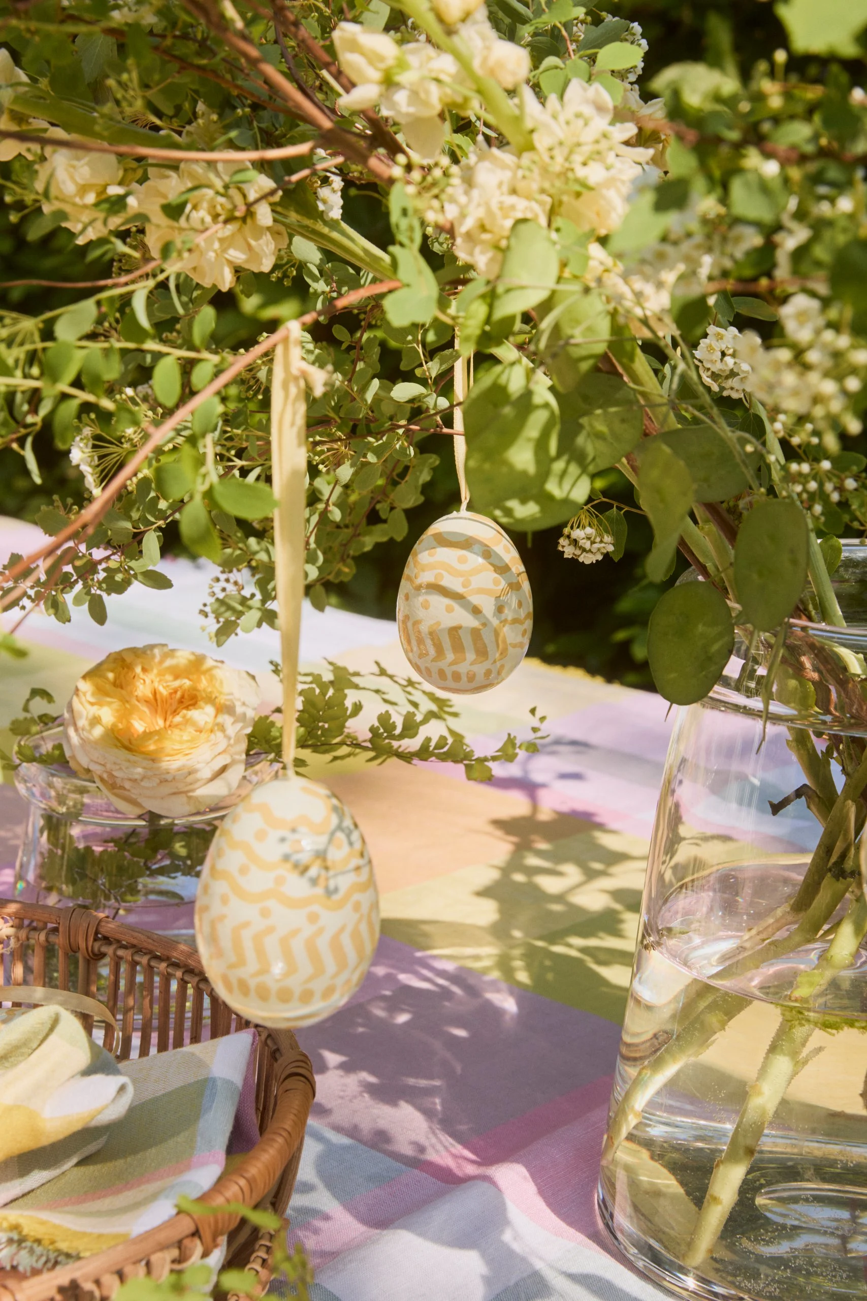 The Easter Egg Easter pendants from Lexington hang from an Easter bush, which is placed outside on a colourfully decorated table.