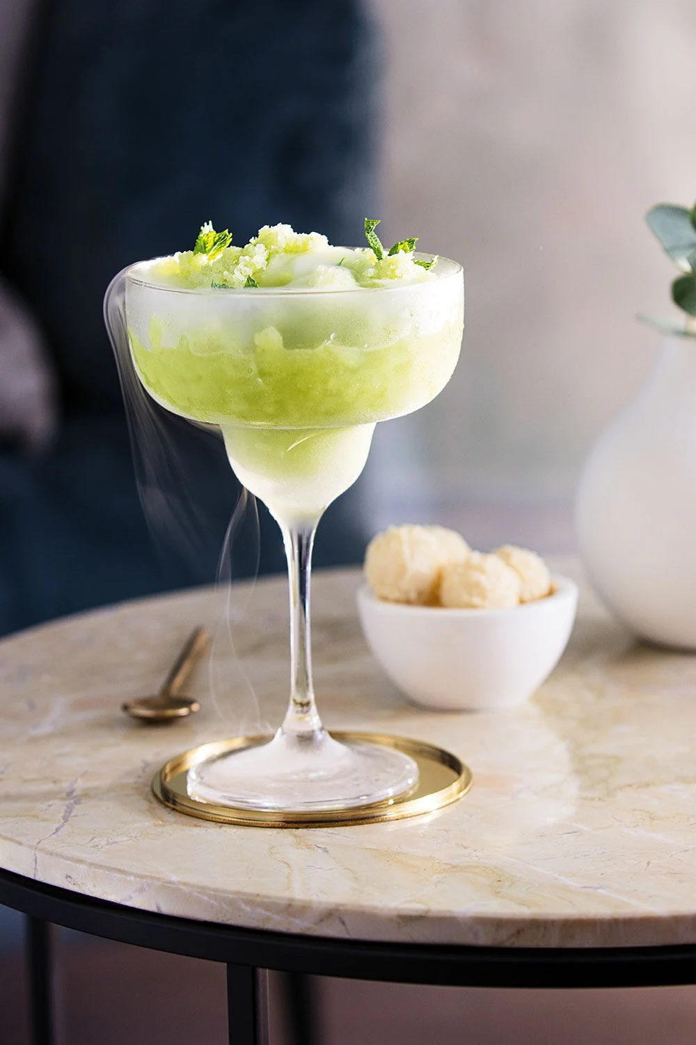 Frozen green drink in a margarita glass with mint garnish, on a marble table with a golden coaster.