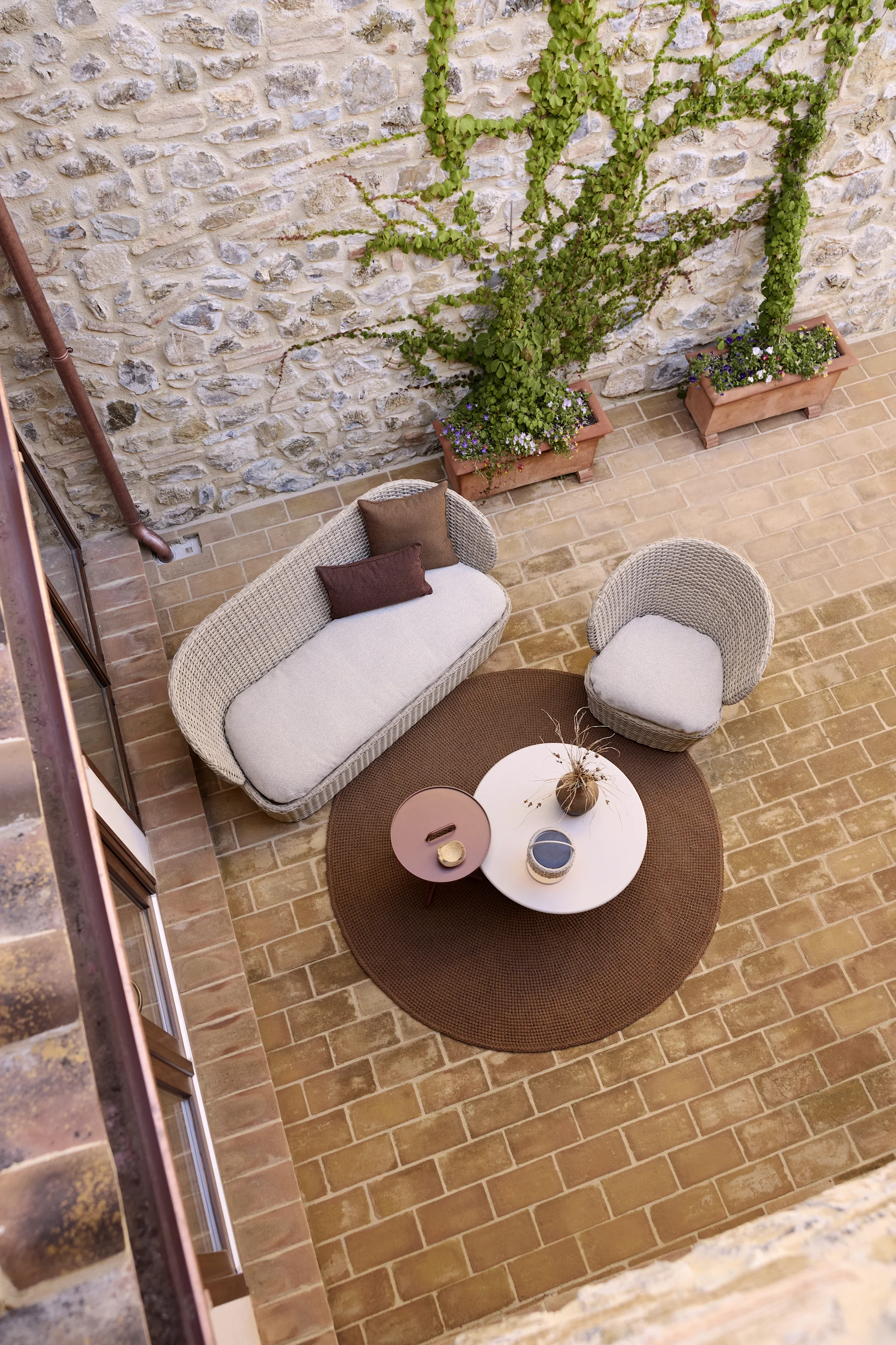 Overhead view of a patio with a woven sofa, armchair, two round coffee tables on a brown rug, and ivy-covered stone walls.