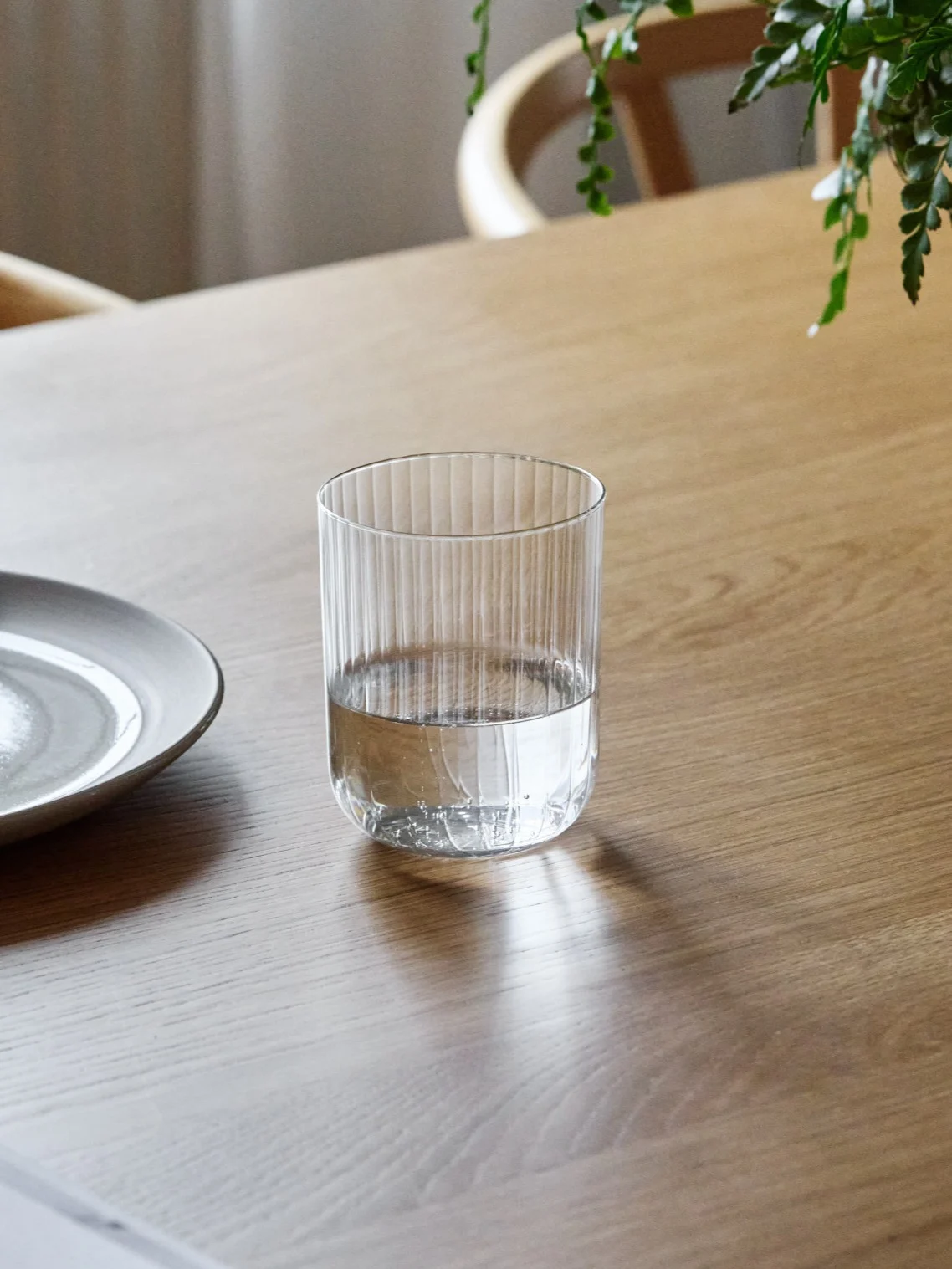 A ribbed glass tumbler, half-filled with water, sits on a wooden table next to a ceramic plate.
