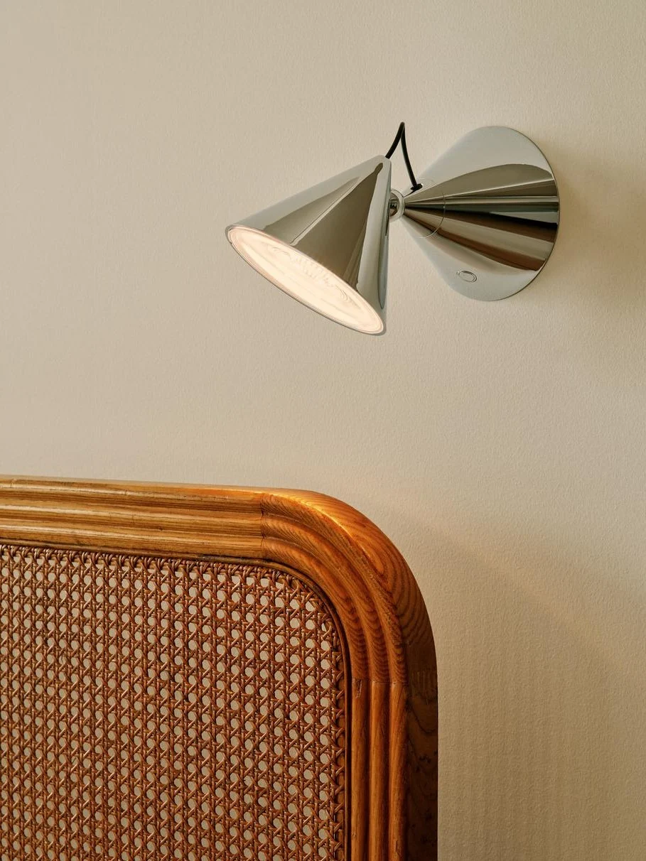 Modern chrome wall lamp with conical shade illuminating a light wall above a woven cane and wood headboard.