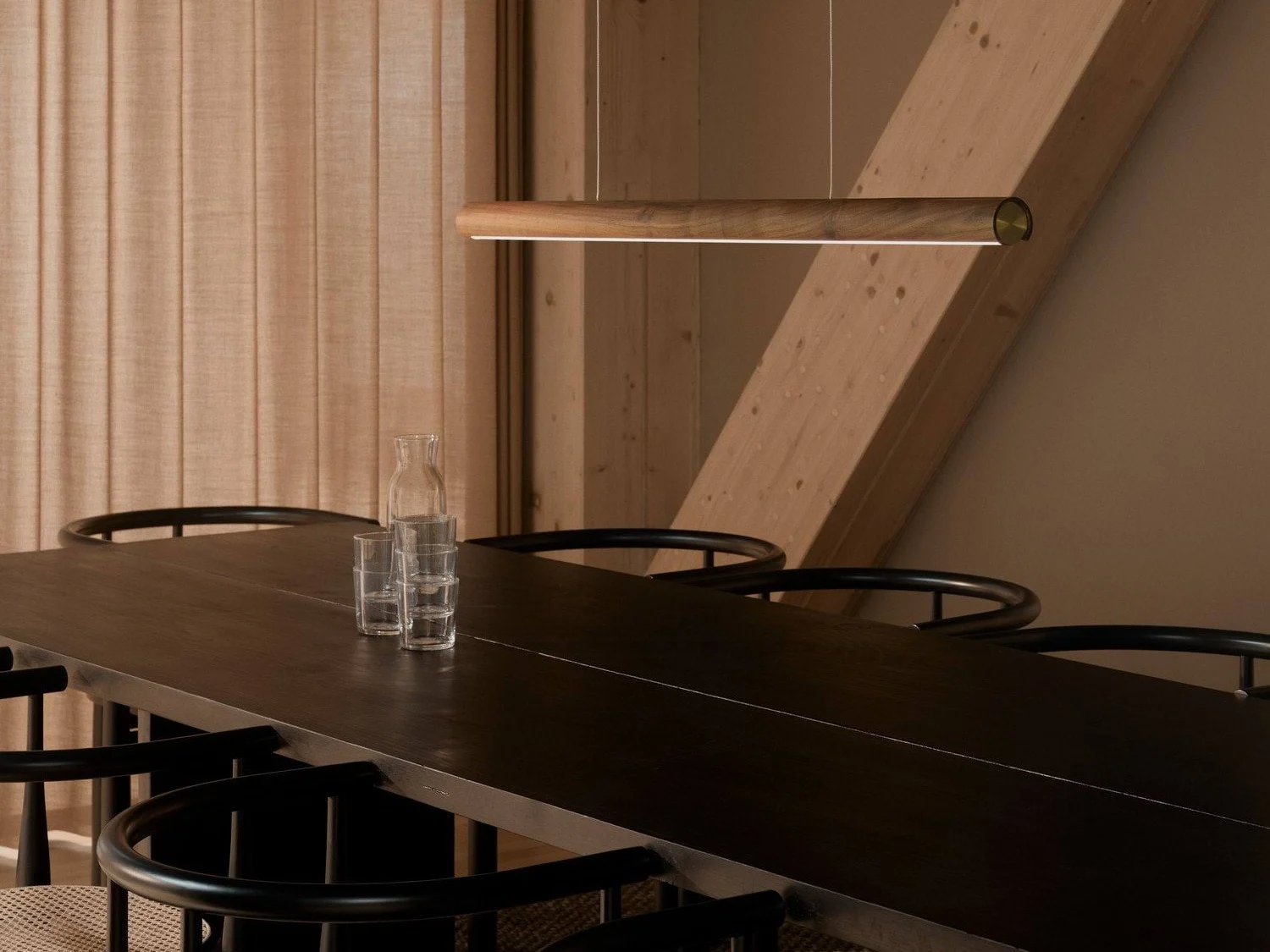 A modern dining room with a long dark wooden table, black chairs, and a sleek wooden pendant light. A glass carafe and three glasses are on the table.