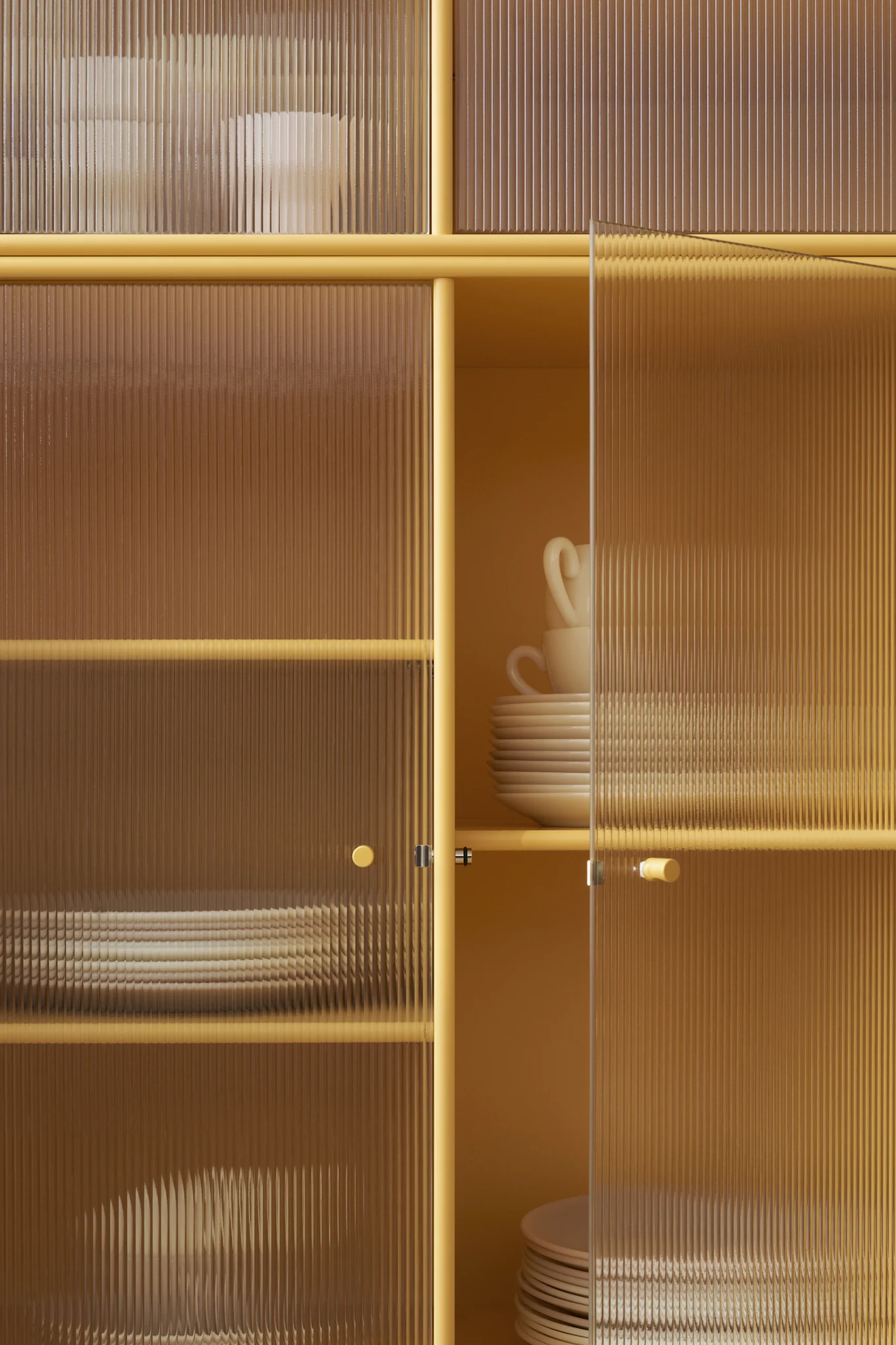 Mustard yellow cabinet with fluted glass doors, revealing stacked white plates and cups inside. One door is ajar.