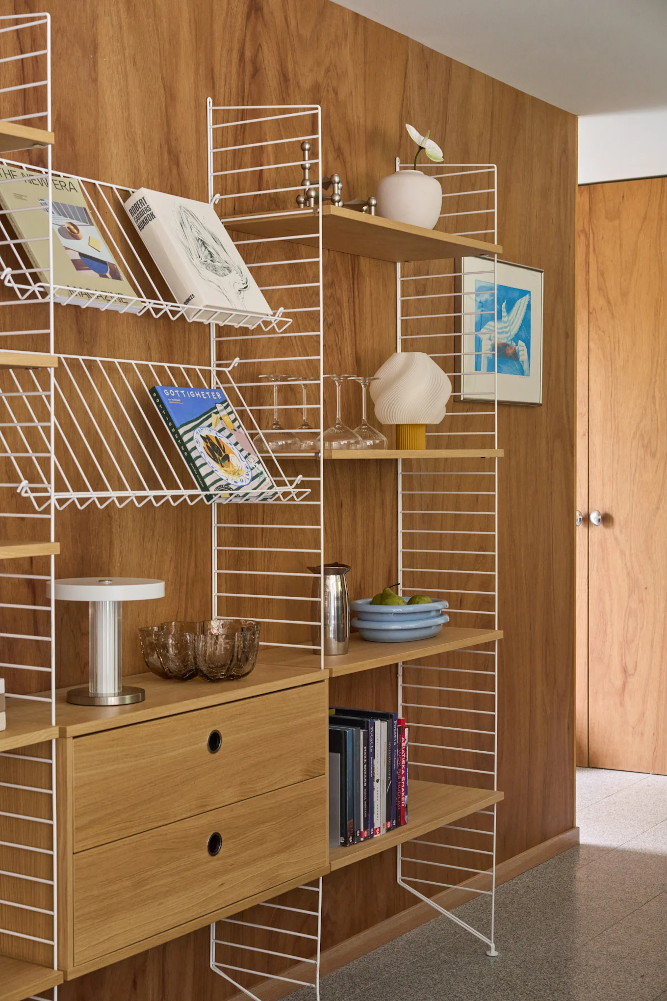 White wire shelving unit with wooden shelves and drawers filled with books, decor, glassware, and a framed artwork against a wood-paneled wall.