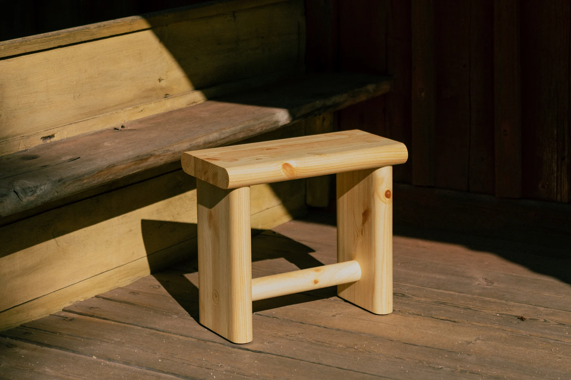 A light wooden stool sits on a wooden floor, partially illuminated by sunlight, next to wooden steps and a dark wooden wall.