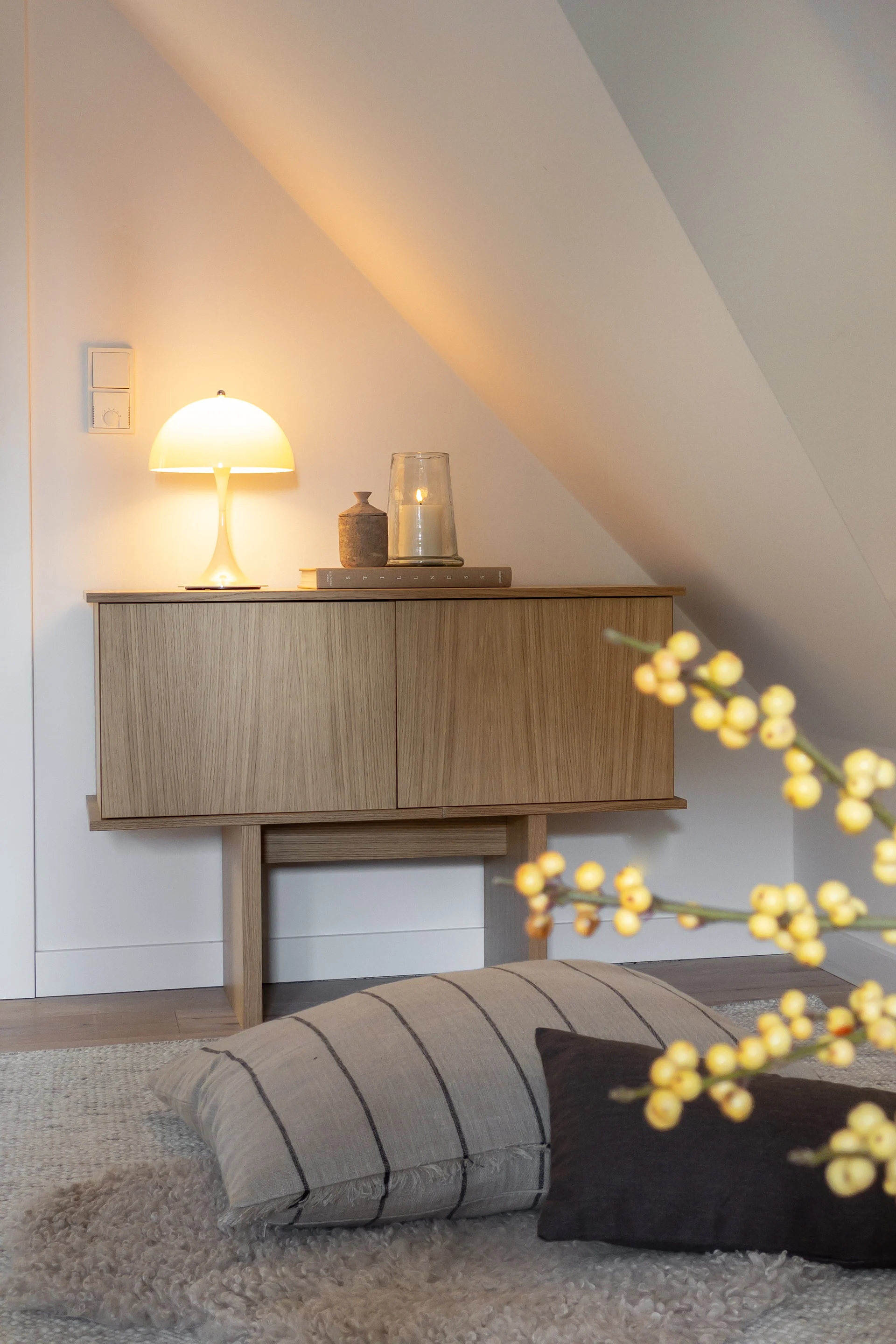 A wooden cabinet with a glowing mushroom lamp and a lit candle, set against a white, sloped wall in a cozy room.