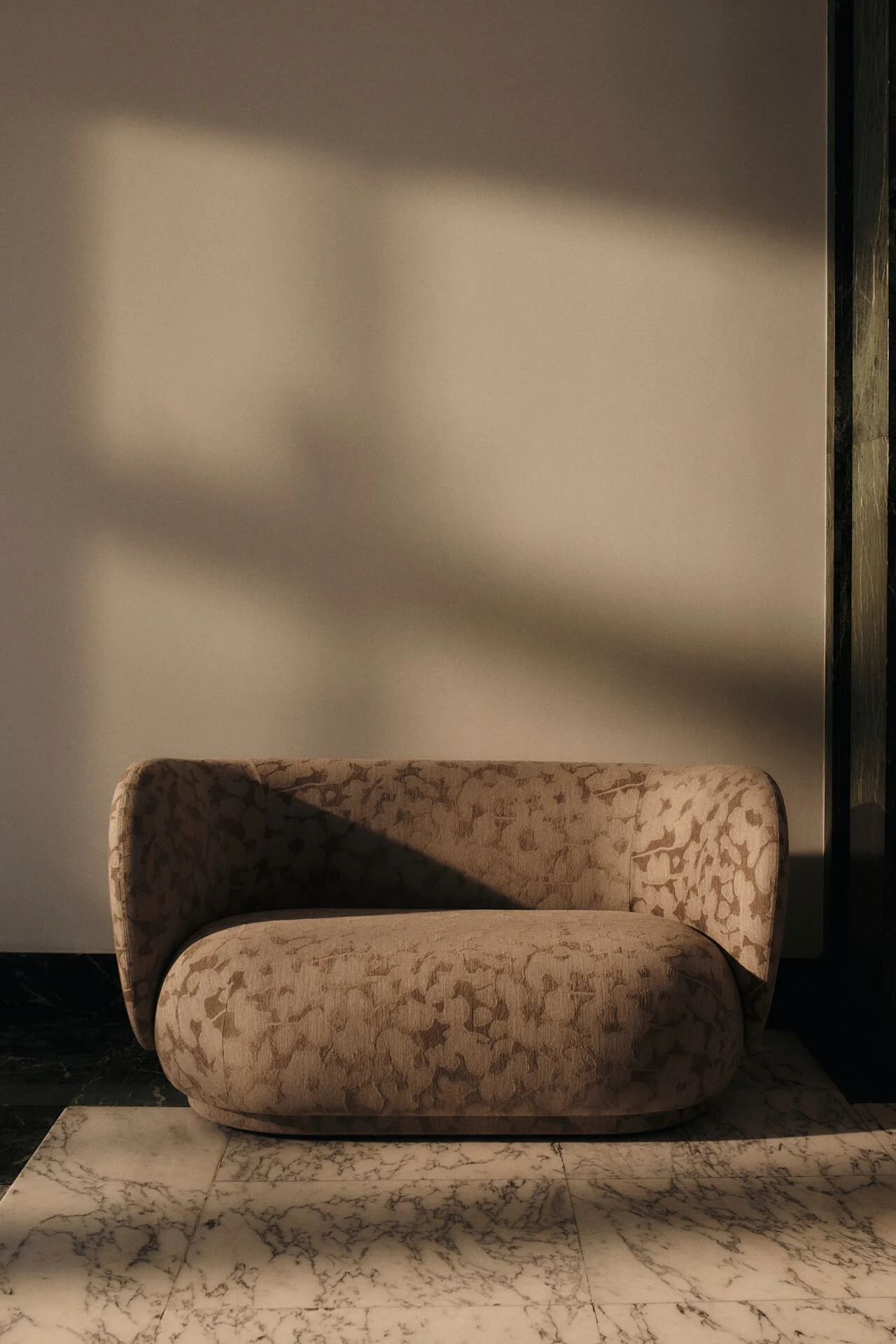 Rounded beige armchair with a subtle patterned fabric on a white marble floor, with dramatic shadows cast by warm light.