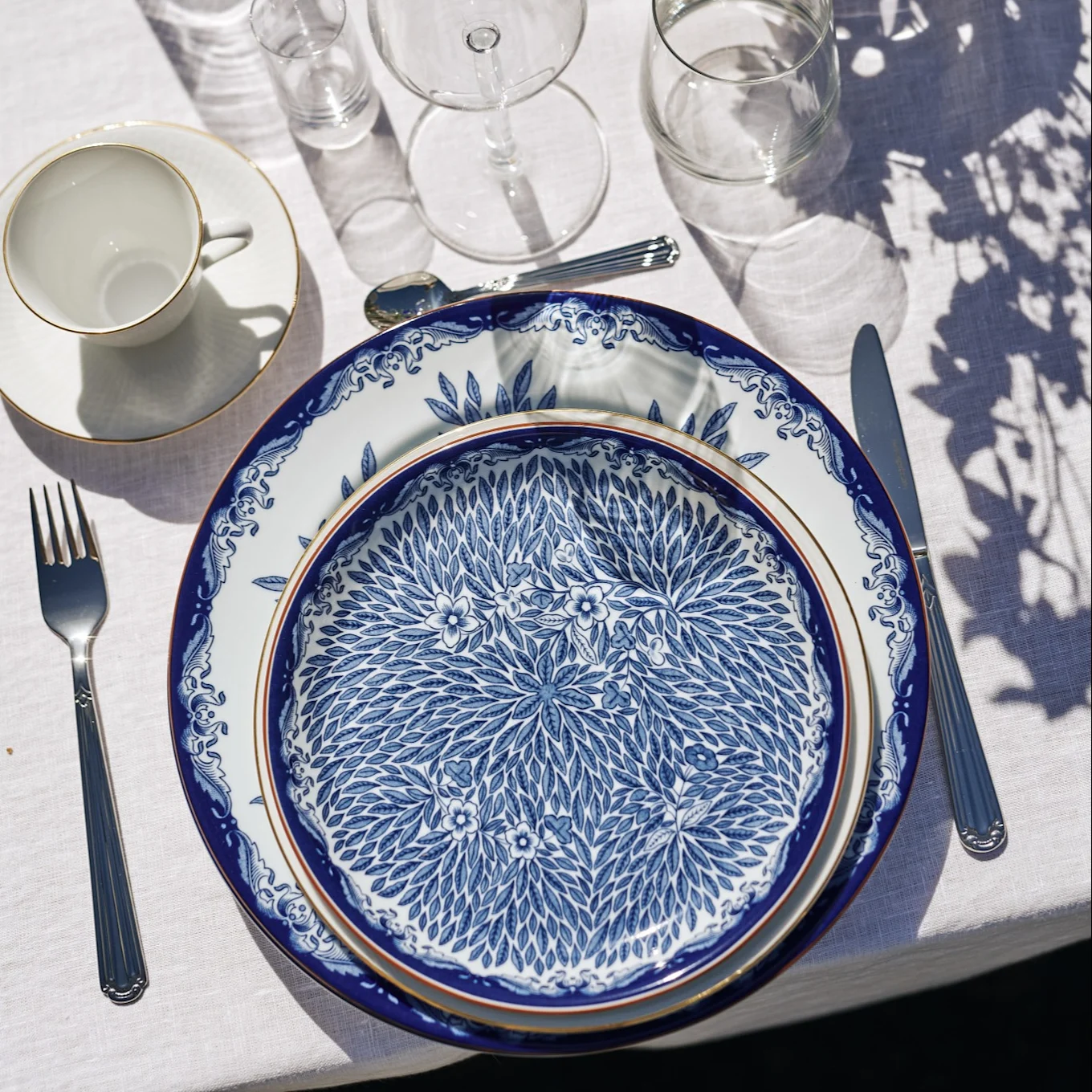 A table setting with blue and white patterned plates, cutlery, a teacup, and glasses on a white tablecloth under dappled sunlight.