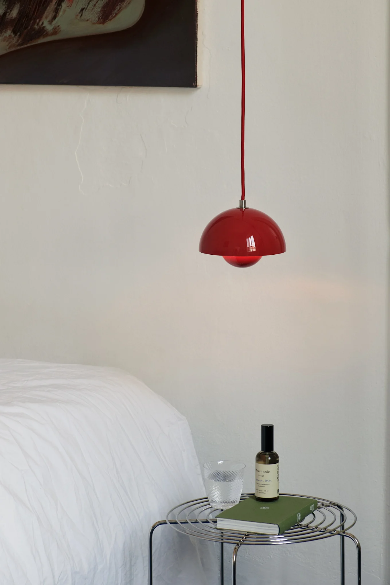Red pendant light hangs above a chrome wire side table with a glass of water, a green book, and a spray bottle in a bedroom.