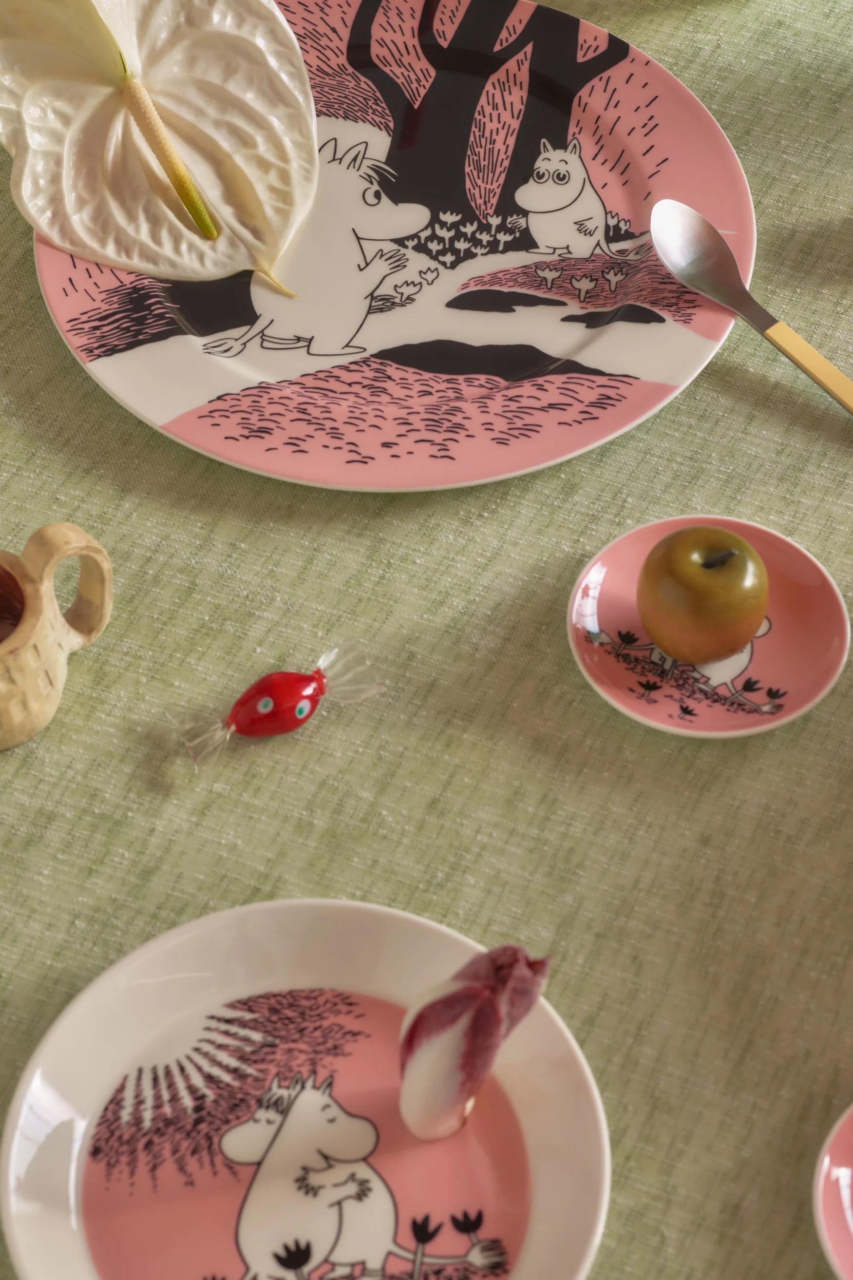 Table set with pink Moomin plates, anthurium flower, spoon, green apple, red candy, and a wooden cup on a green tablecloth.