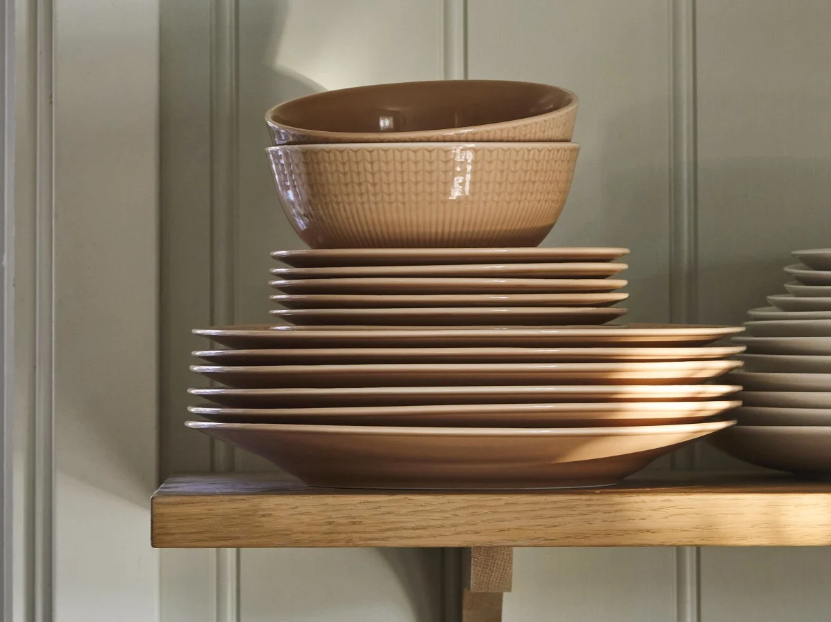 Stacked light brown ceramic plates and two ribbed bowls on a wooden shelf.