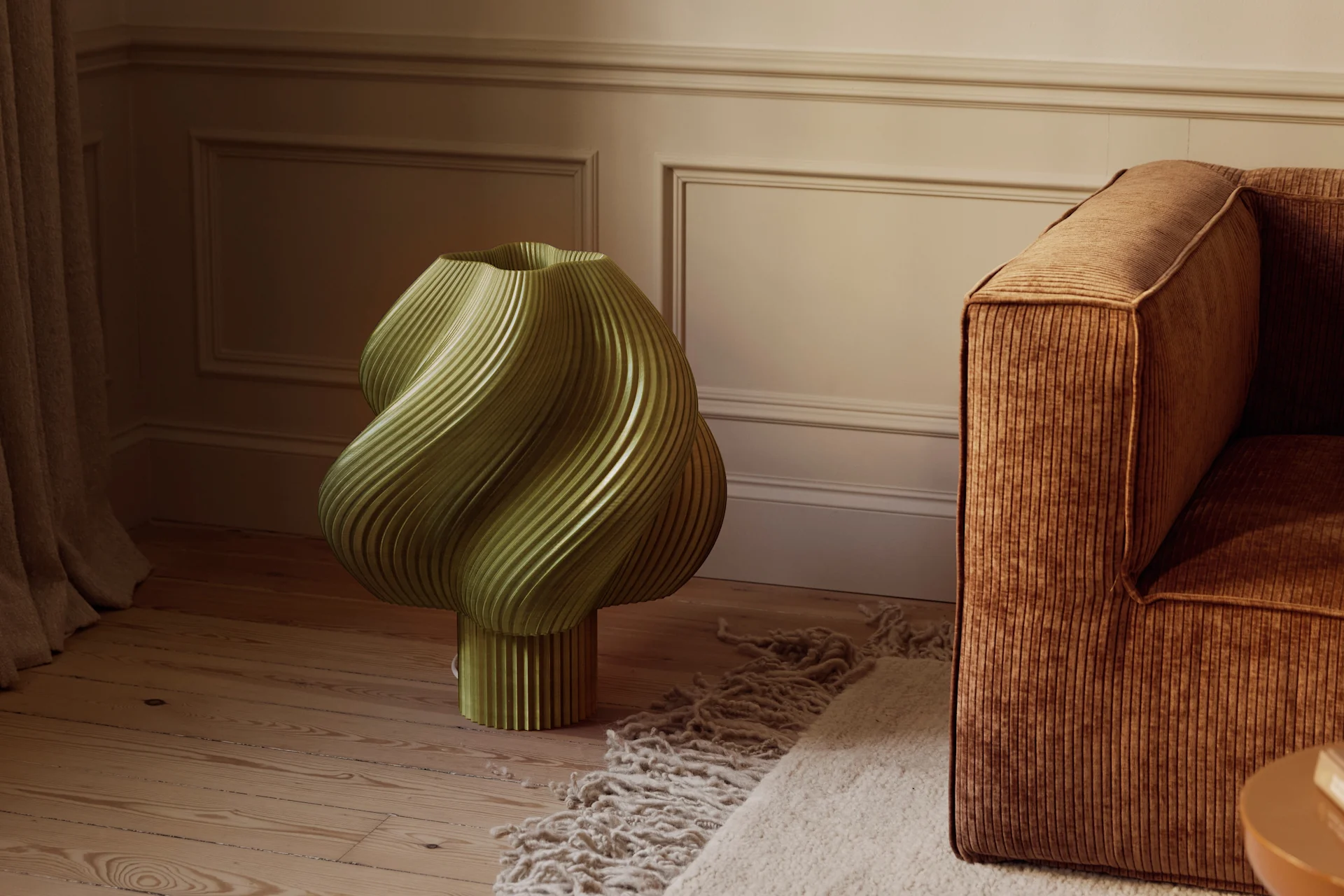 An olive green, sculptural, fluted object on a wooden floor next to a brown corduroy sofa and textured rug in a room with molded walls.