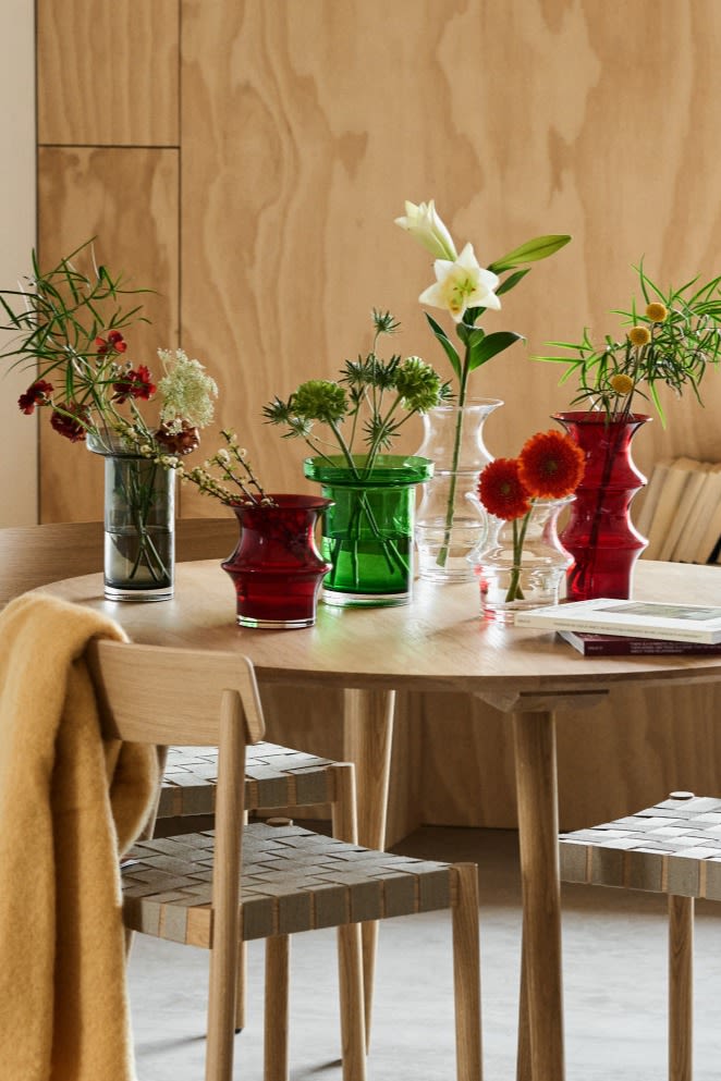 A collection of Kosta Boda vases in clear, red and green glass stand on a dining room table with fresh cut flowers. 