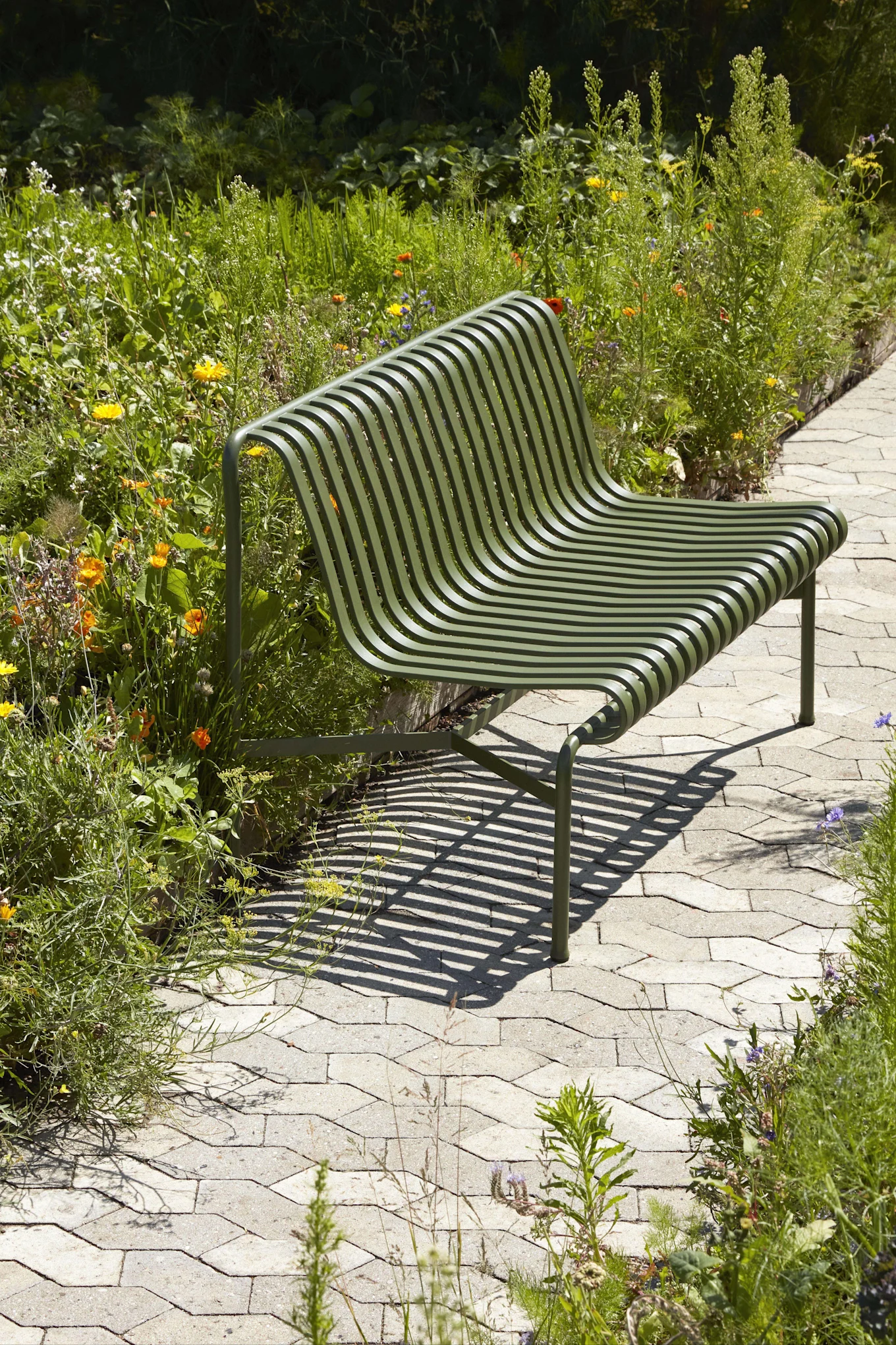 Dark green slatted park bench with a curved back on a paved path, surrounded by lush garden foliage and colorful flowers.