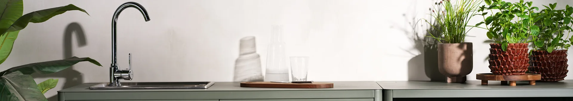 A modern kitchen counter with a sink, faucet, glass bottle, drinking glass, and several potted green plants against a white wall.