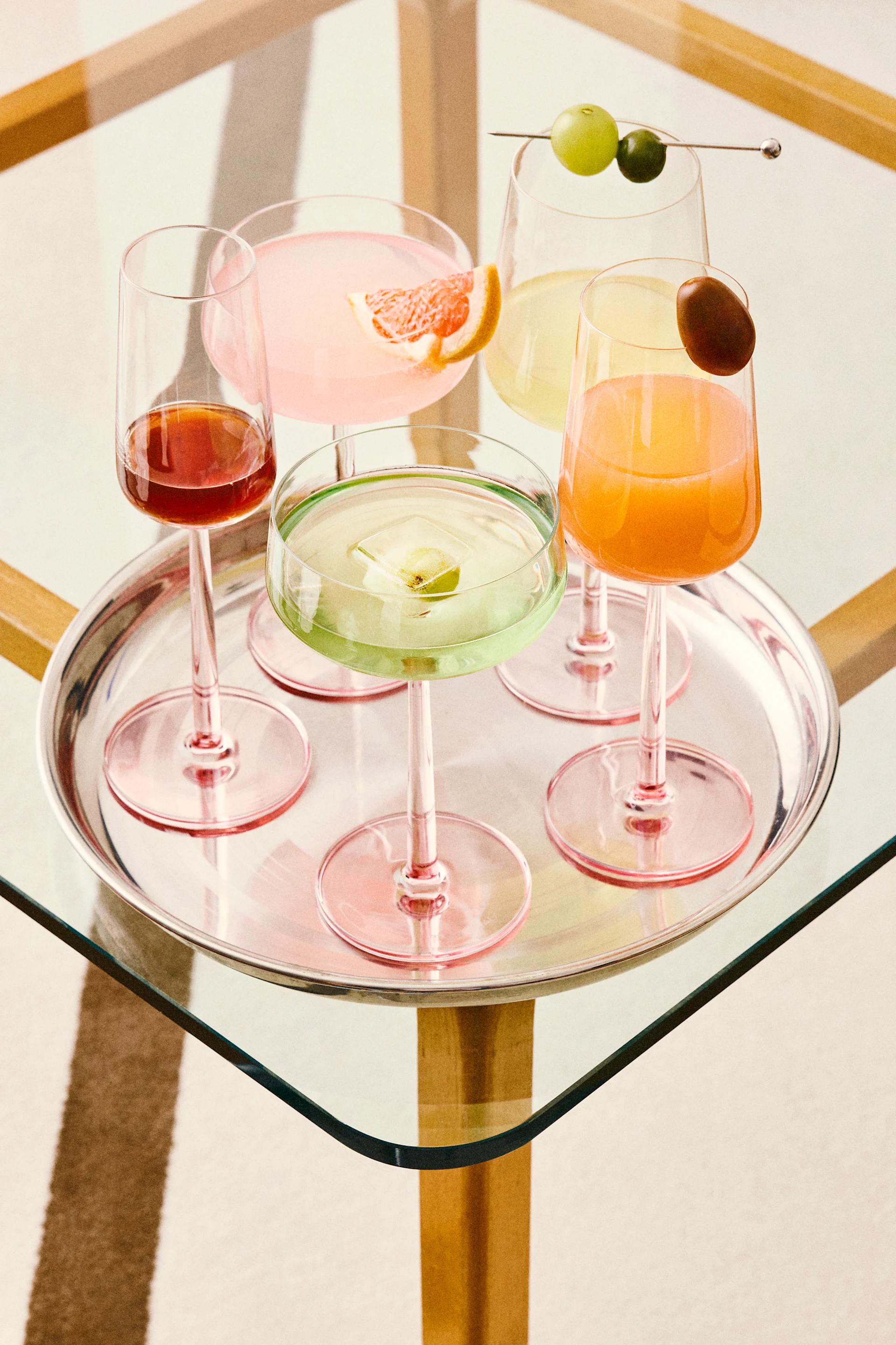 Five colorful cocktails on a silver tray, including pink, green, orange, and red drinks with various garnishes, on a glass table.