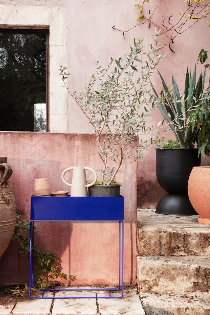Metal outdoor plant box in cobalt blue from Ferm Living, here filled with terracotta pots, a vase and a large plant with green leaves.