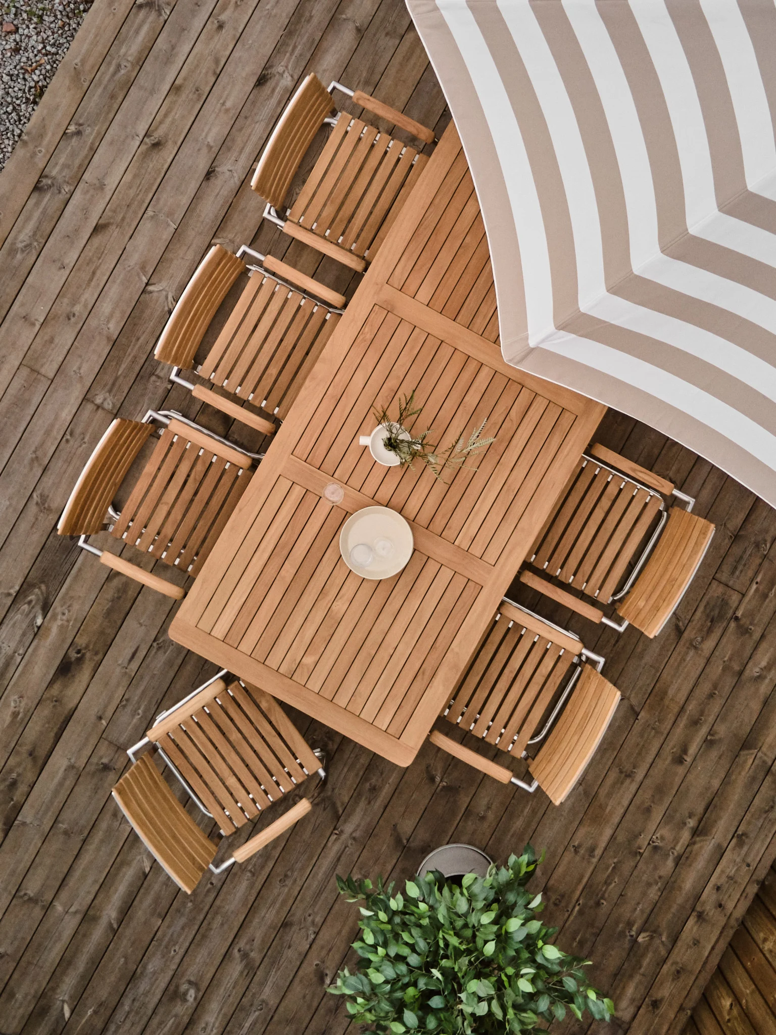 Overhead of a wooden patio dining set with a rectangular table, eight slatted chairs, and a beige-white striped umbrella.