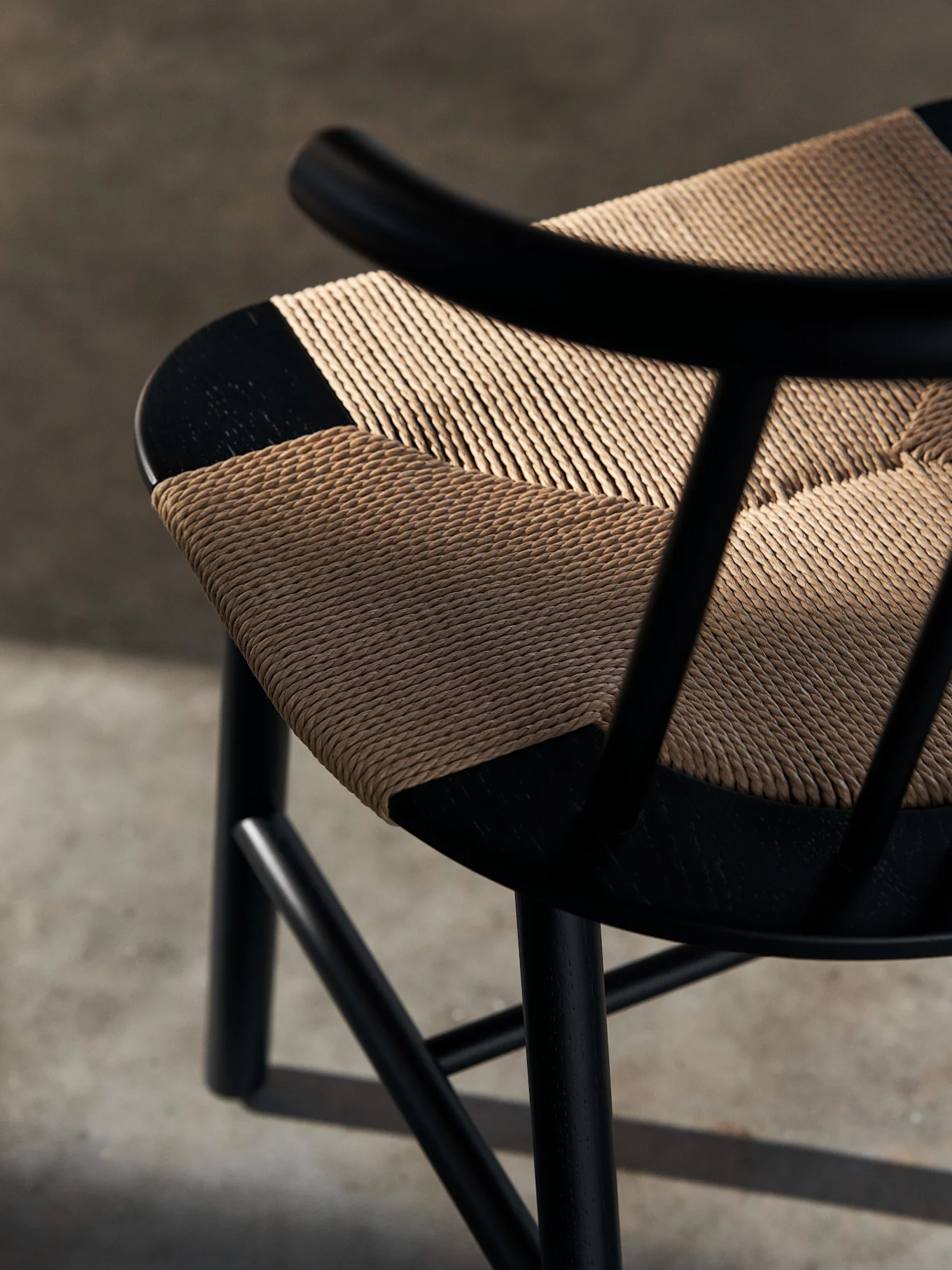 Close-up of a black wooden chair with a light tan, diagonally woven seat. Shadows fall across the textured seat.