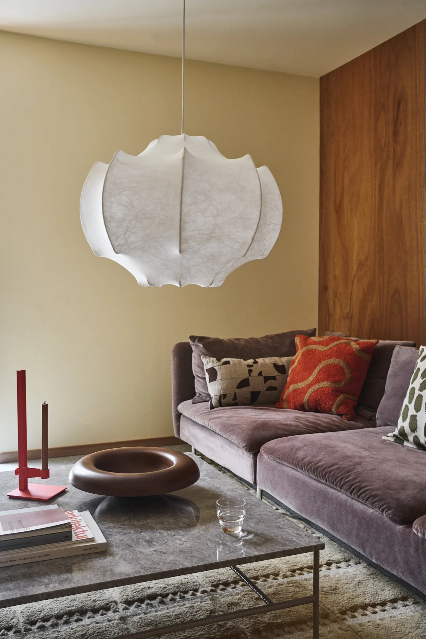 A modern living room with a white sculptural pendant lamp, a purple velvet sectional sofa with colorful pillows, and a marble coffee table.