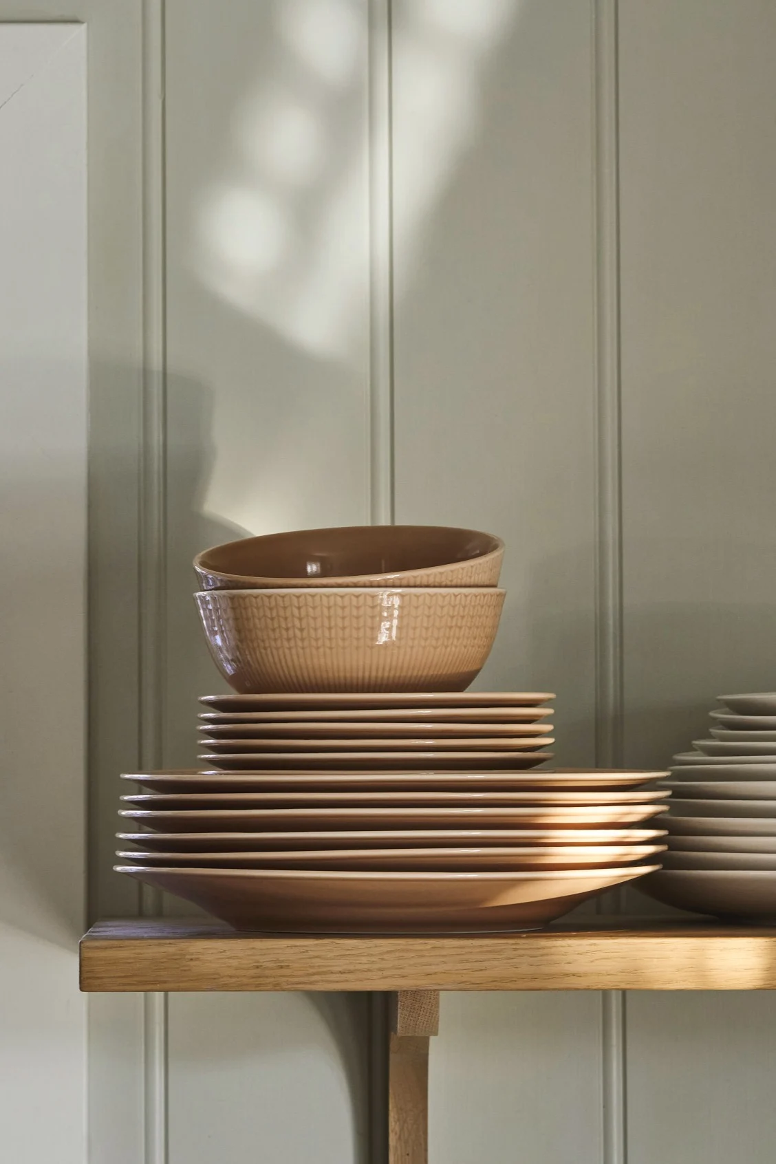 Stack of light brown ceramic plates and bowls with a textured pattern on a wooden shelf against a paneled green wall.