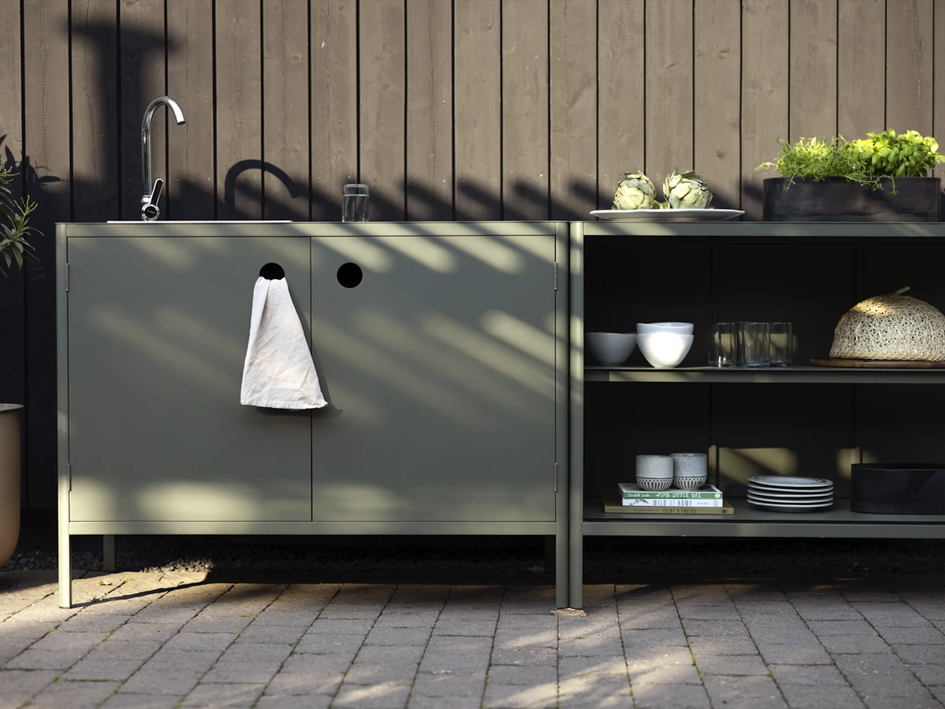 Outdoor kitchen with dark green cabinets, a sink, faucet, and shelves holding artichokes, herbs, and dinnerware.