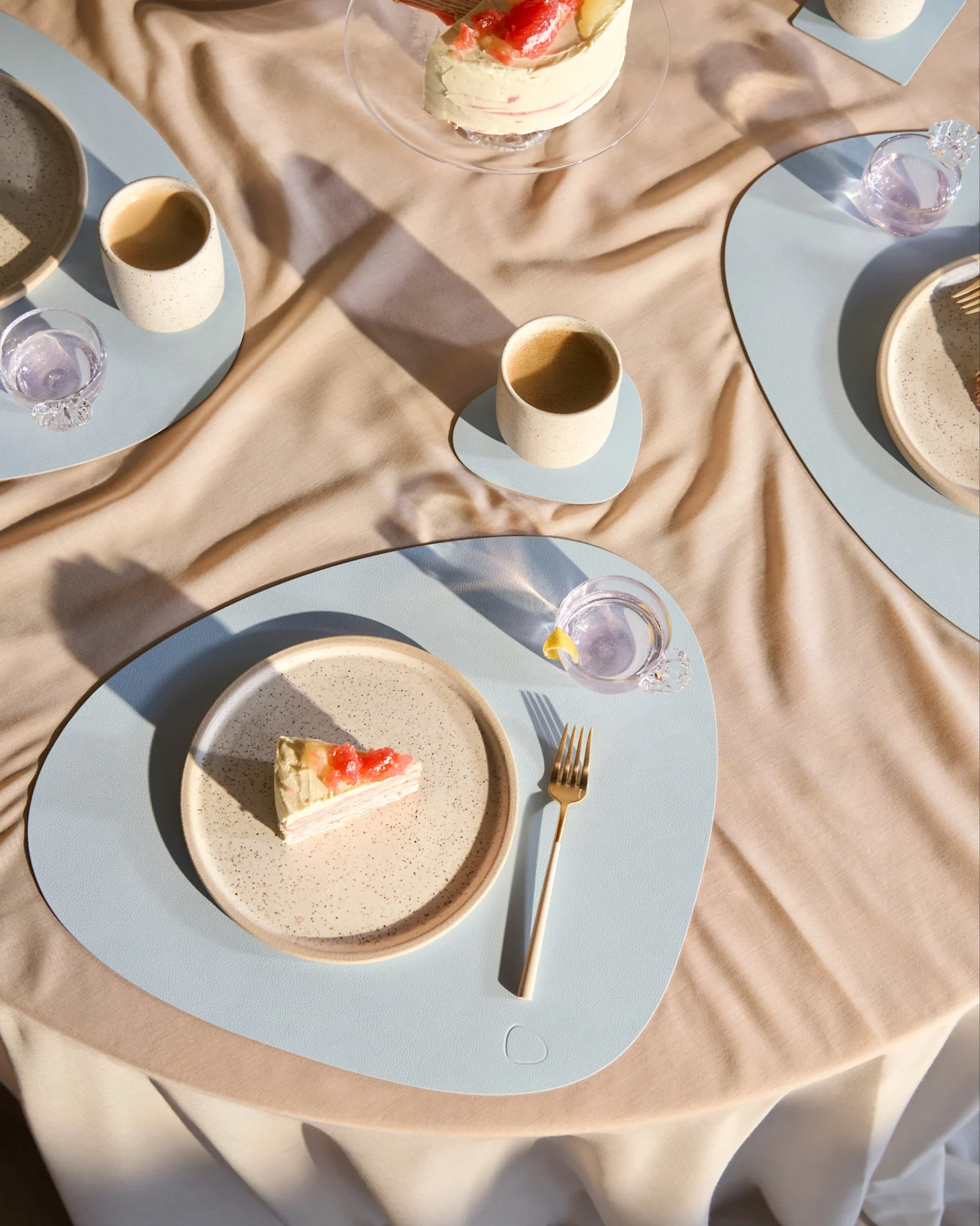 Table set with a crepe cake topped with grapefruit and pineapple, a slice on a plate with a gold fork, cups on light blue placemats.