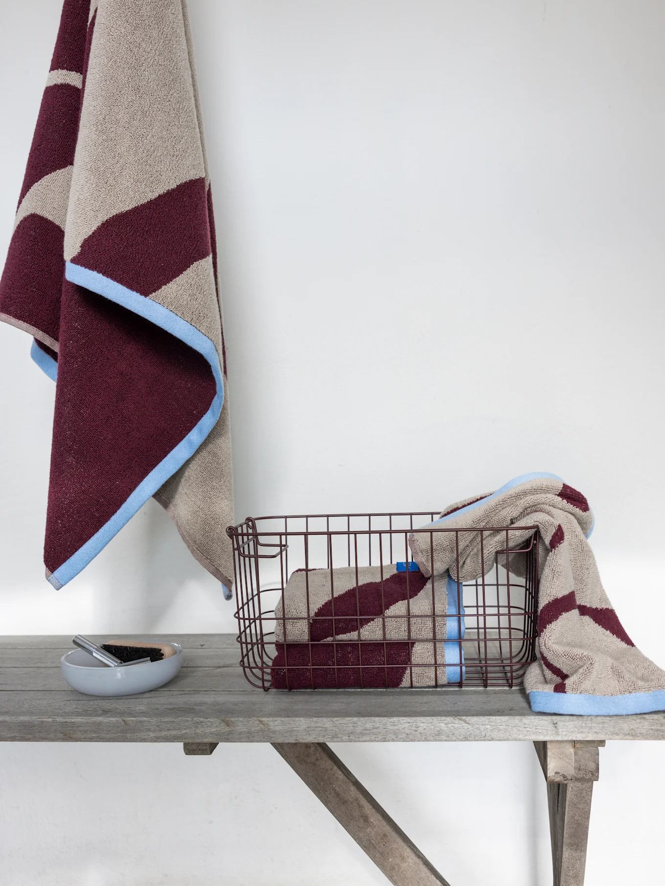 Two maroon and beige striped towels with light blue borders. One hangs, another is in a dark wire basket on a wooden shelf.