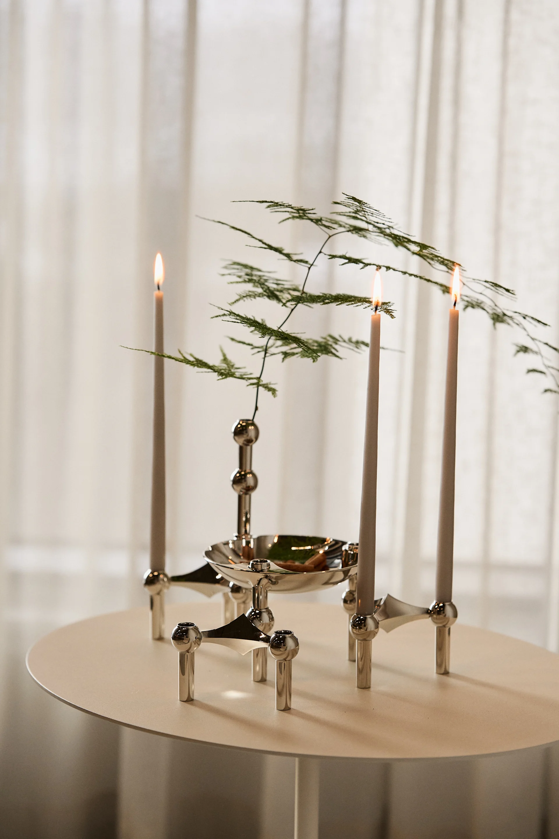 Three lit candles in a modular silver candlestick with green foliage, on a light round table.