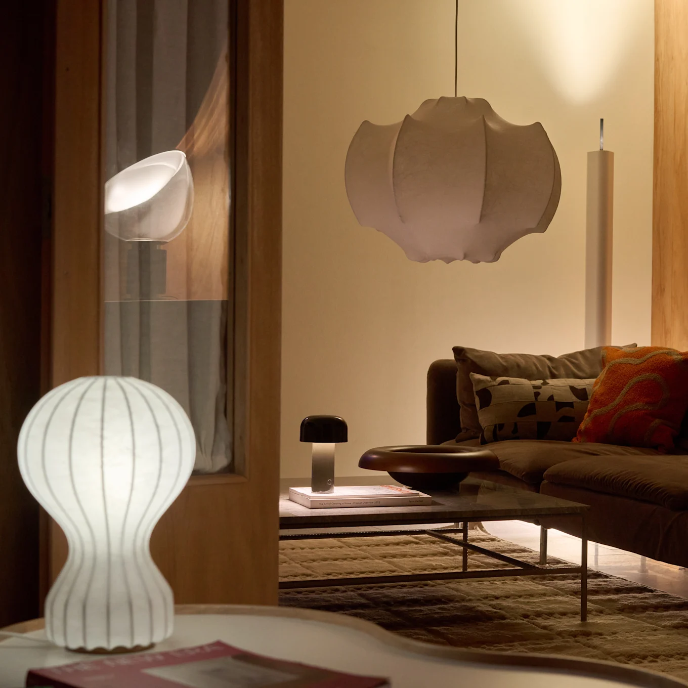 A living room with a brown sofa and coffee table, illuminated by a white ribbed table lamp, a dark mushroom lamp, and a large pleated pendant lamp, creating a warm glow.