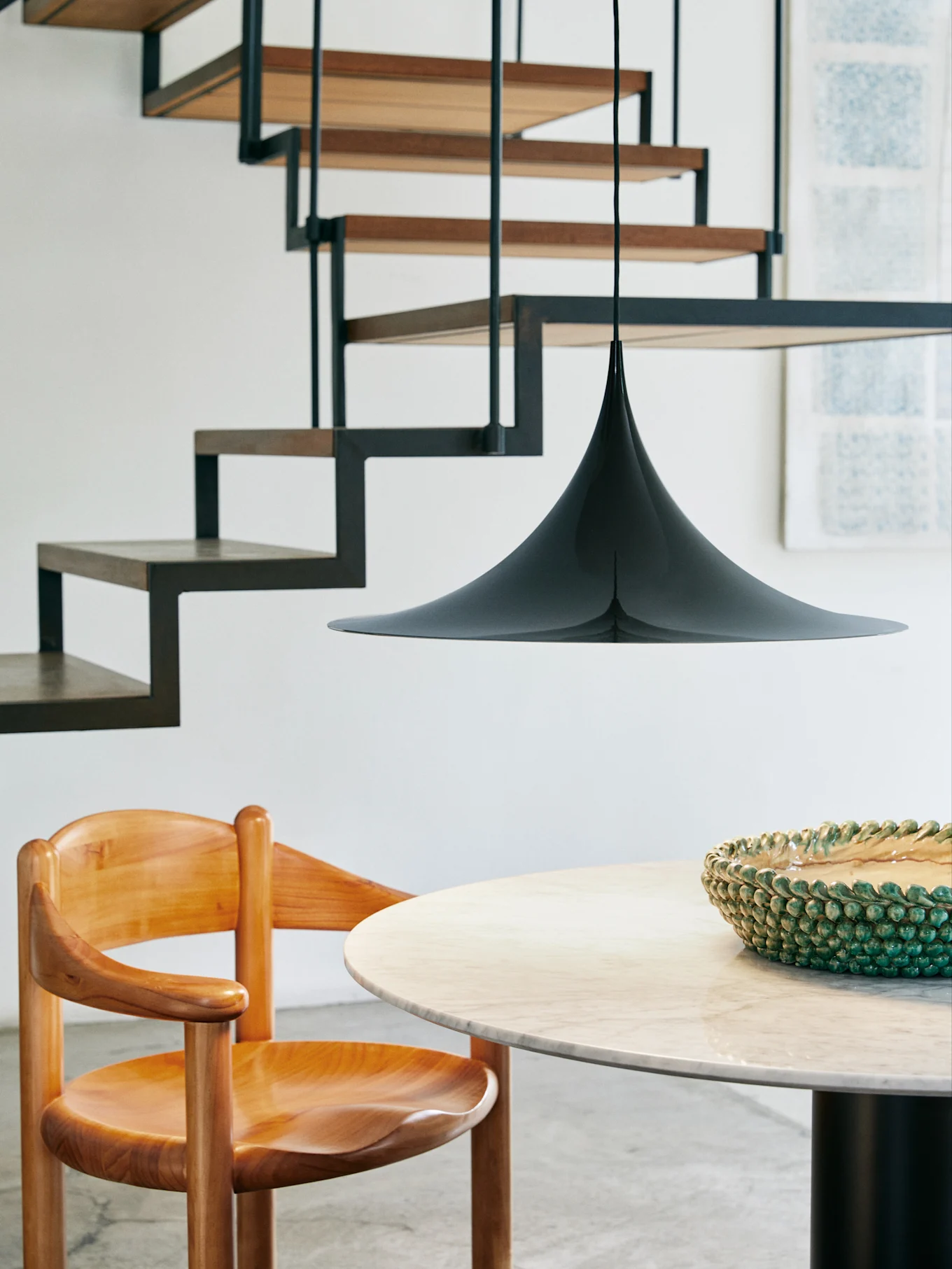 A black cone-shaped pendant lamp hangs over a round marble table and a wooden chair, with a modern staircase in the background.