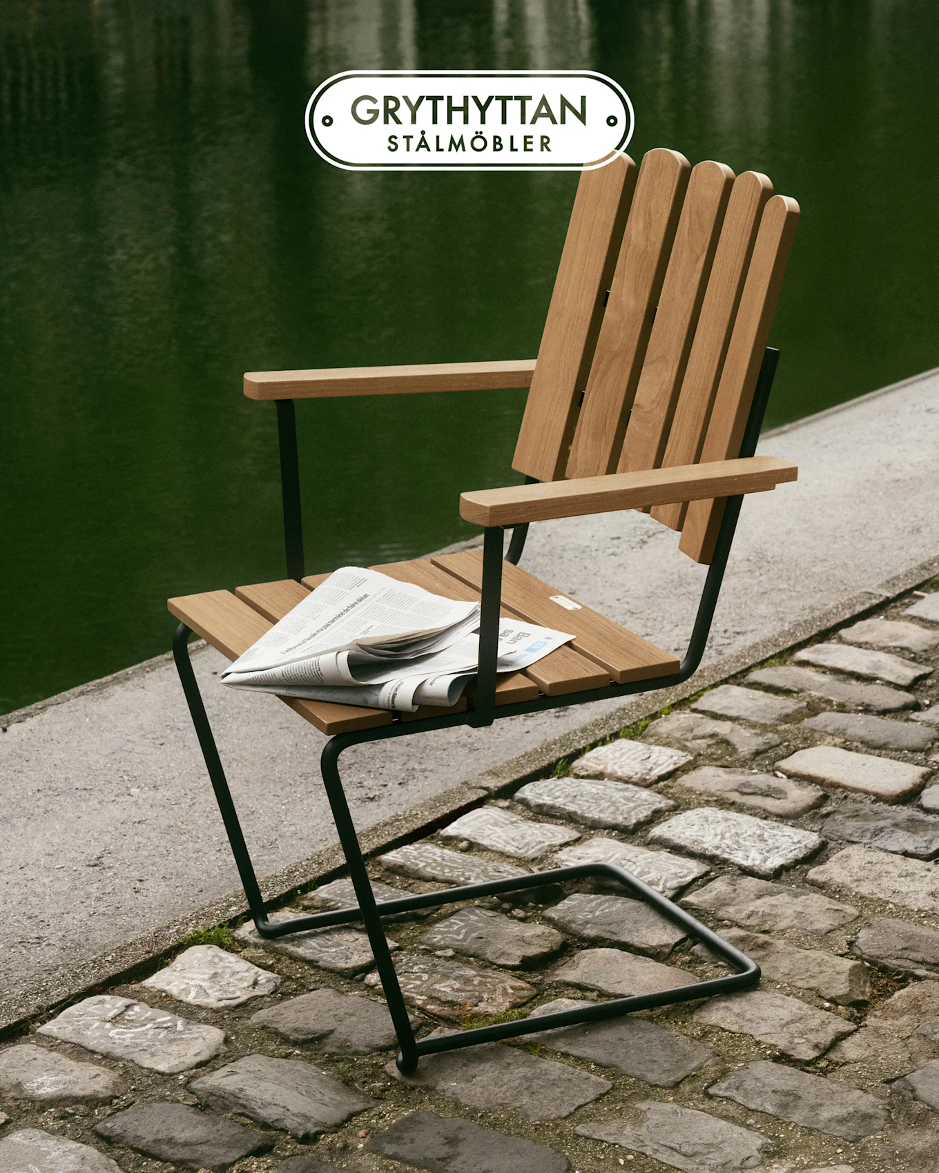 Wooden slat chair with black metal frame on a cobbled path by a green body of water, newspaper on seat.