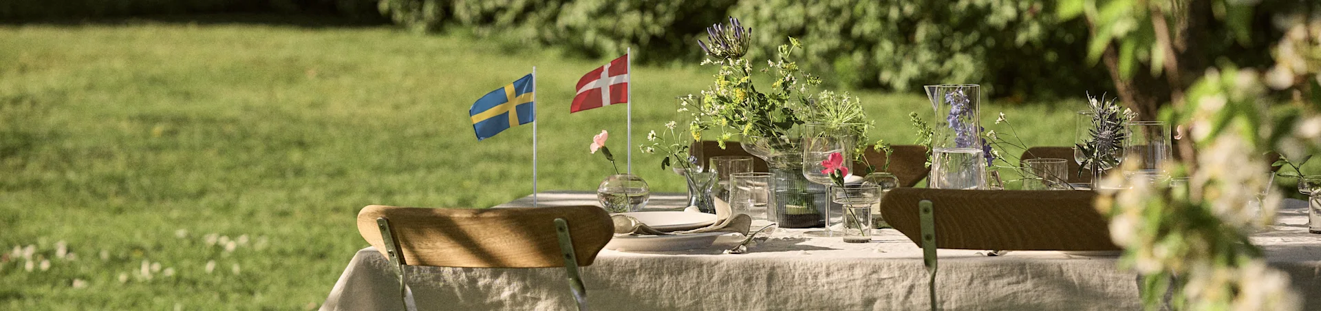 Outdoor dining table set with white linen, glassware, floral arrangements, and Swedish and Danish flags in a sunny garden.