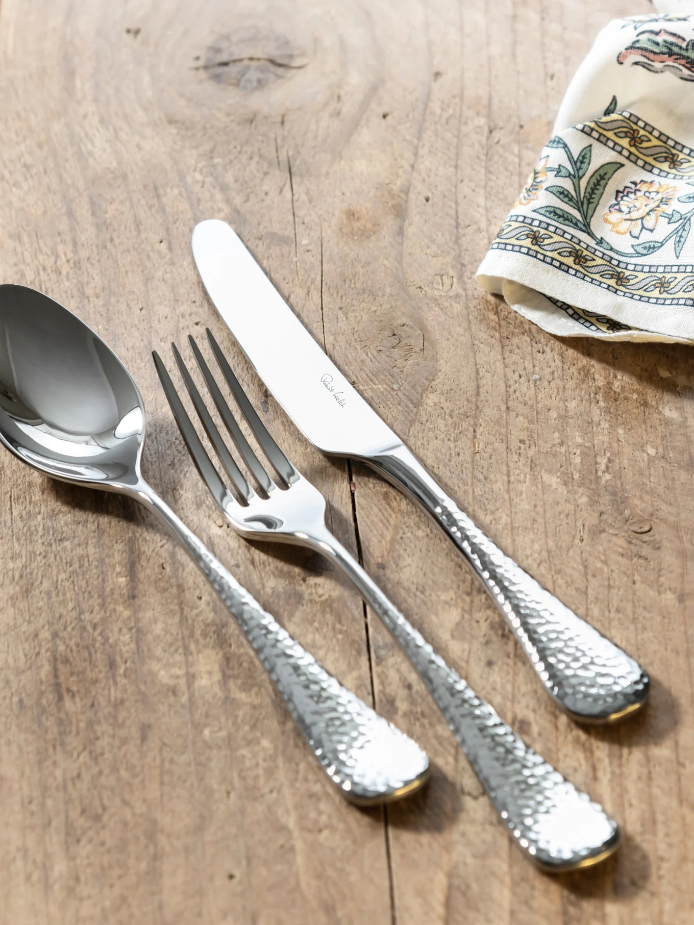 A set of silver-colored cutlery with hammered handles on a wooden table, with a patterned napkin. The knife is inscribed 'Robert Welch'.
