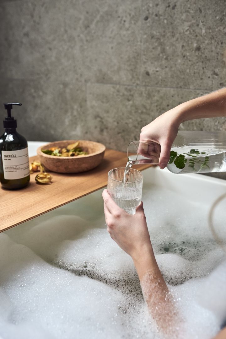Hands pour water into a ripple glass from Ferm Living as part of a home spa night with a warm bubble bath. 
