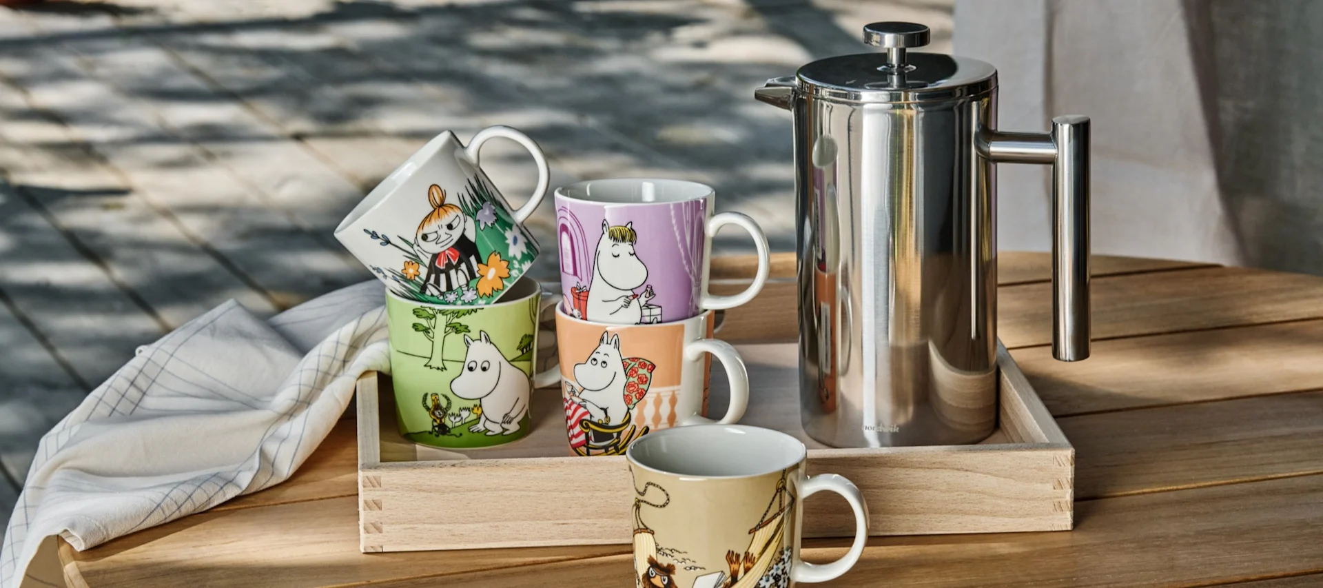 Various Moomin cups from Moomin Arabia stand on a table outside, next to a stamping jug.