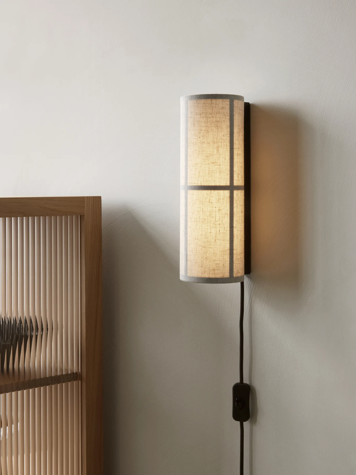 Cylindrical wall lamp with a translucent fabric shade, mounted on a light wall next to a wooden slatted screen.