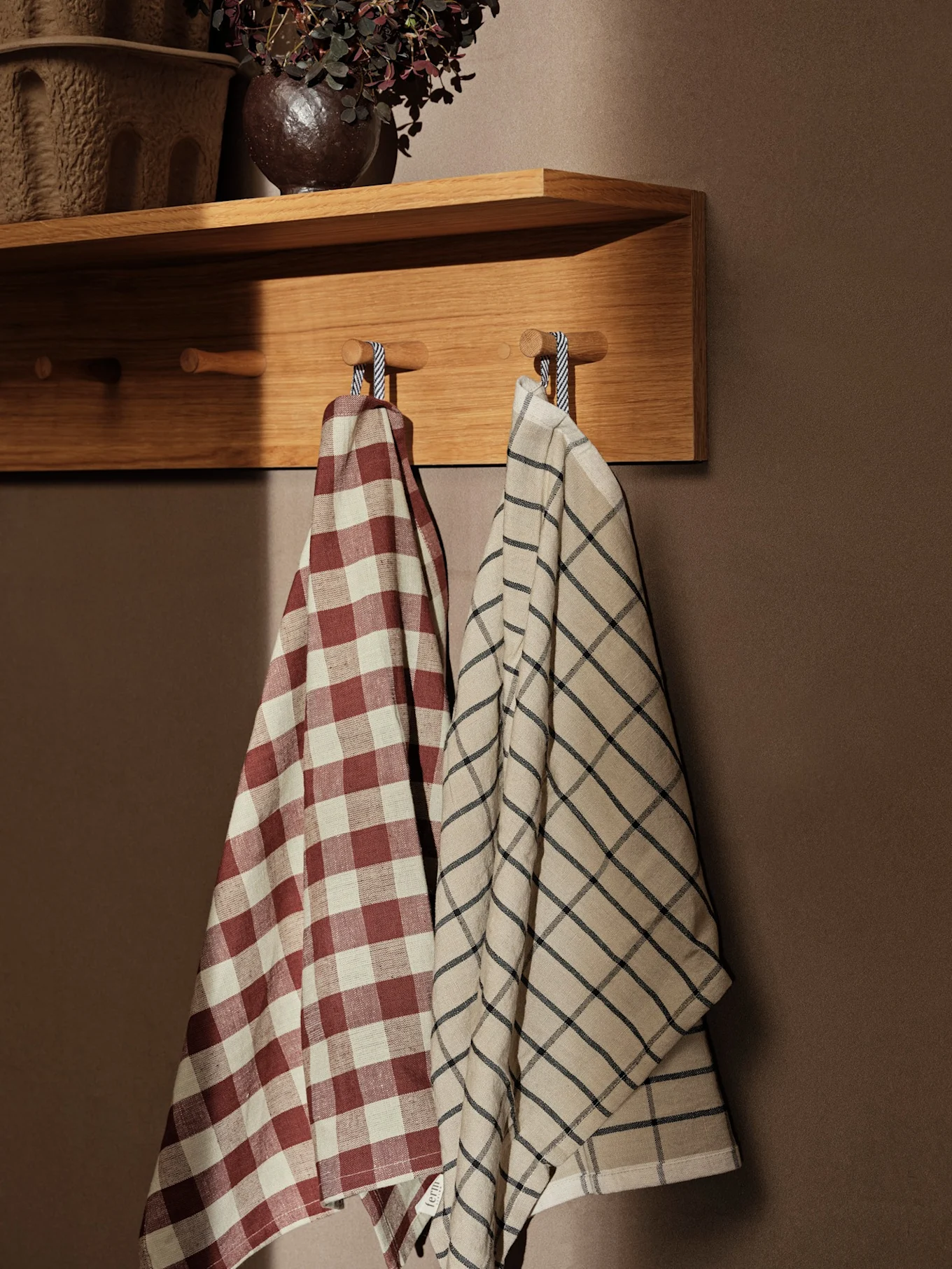 Two checkered kitchen towels, one red and cream, the other cream with a black grid, hanging on wooden pegs below a shelf.