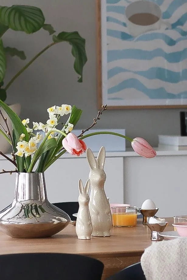 A table set for spring breakfast with a silver vase of daffodils and tulips, two ceramic bunny figurines, orange juice, and an egg.