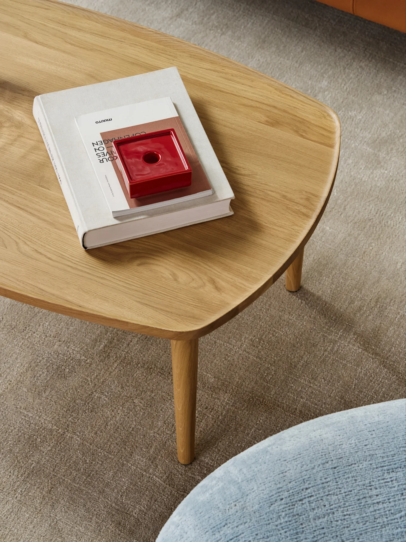 A light wood coffee table with rounded edges, holding a stack of books and a red ceramic object, on a beige carpet.